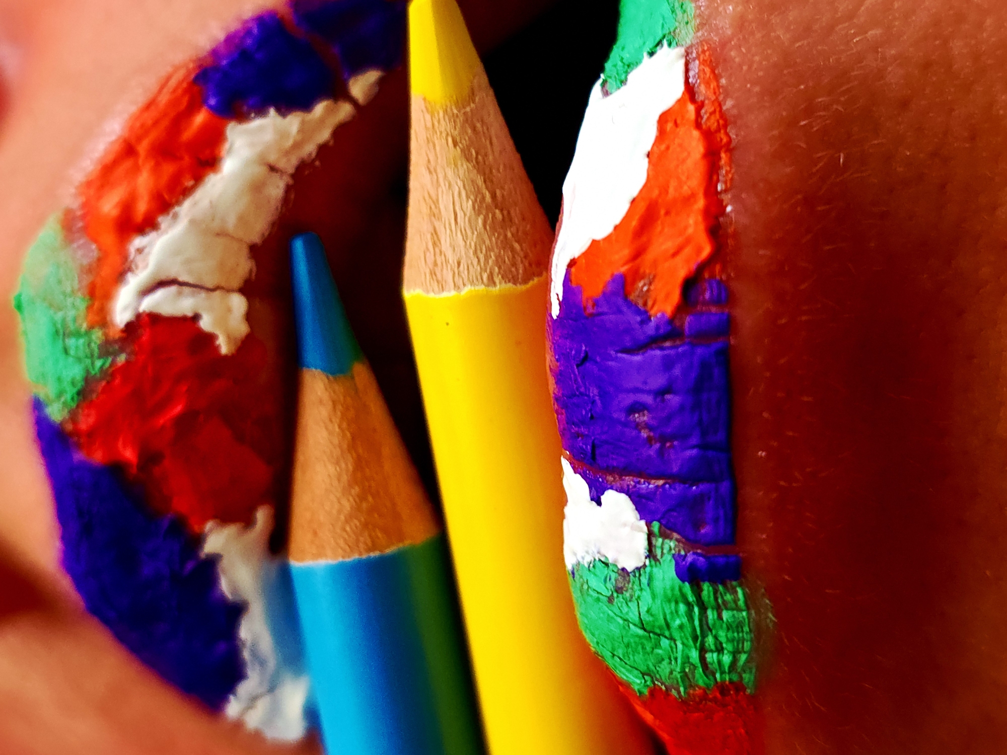 The image depicts a close-up of colored pencils held against lips painted with various colors. The pencils are blue, yellow, and brown. The paint on the lips includes green, red, white, and purple. The location and the identities of any people involved are unknown.