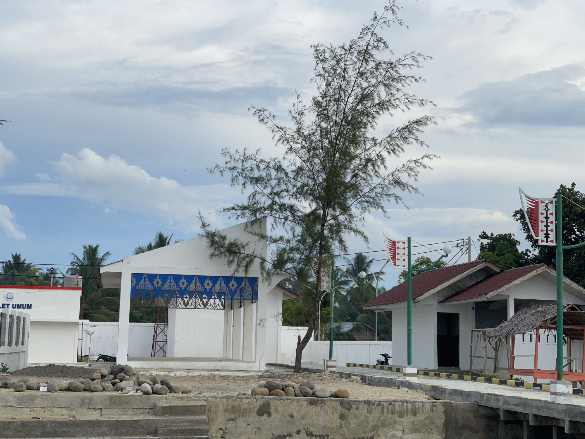 The image depicts an outdoor scene in Lancok, Indonesia, likely during the daytime given the bright overcast sky. The weather appears cloudy. The setting is a coastal or waterfront area with buildings and a pathway.
In the foreground, there is a concrete structure with steps leading up to a sandy area. A pile of smooth, grey stones is scattered on the sandy ground. Beyond this, a white building with an open-air section and decorative blue and gold patterned panels is visible. To the right, another small white building with a red-tiled roof stands. A small, thatched-roof hut is attached to this building. A concrete pathway with yellow and black striped borders runs alongside the buildings. A motorcycle is parked near the pathway.
A tall, slender tree with sparse, needle-like leaves stands in the middle ground. Two flagpoles with distinctive red and white flags are positioned in front of the buildings. Palm trees and other vegetation are visible in the background, suggesting a tropical location.
There is visible text on the white building to the left, reading "LET UMUM". The overall impression is of a developing or recently constructed area, possibly a public facility or tourist spot. The scene is calm and devoid of people.