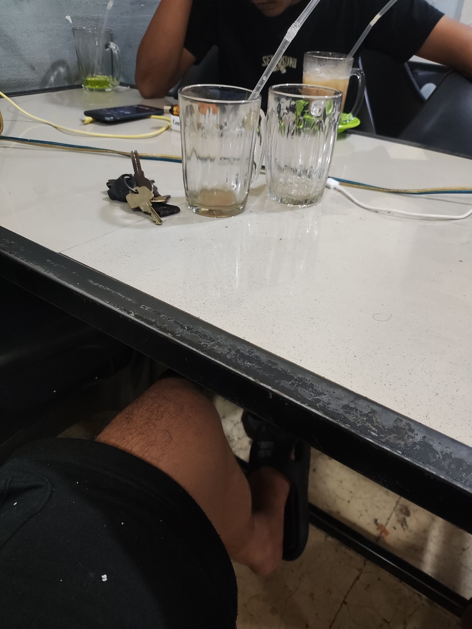 The scene is a table setting in a likely indoor or semi-indoor cafe environment in Lhokseumawe, Indonesia. Two individuals are partially visible. One individual, presumably the photographer, is wearing black shorts and black flip-flops, and is seated at the table. Another individual is wearing a black t-shirt and is seated across the table.
On the table there are three drinking glasses, two of which appear to be empty, and one with a greenish liquid. There is also a drink in a mug with a straw. Other objects on the table include keys, a phone, a cigarette, and various charging cables.
The table itself has a speckled surface and a black metal frame showing signs of wear. The floor is visible beneath the table and has a dirty, textured appearance.