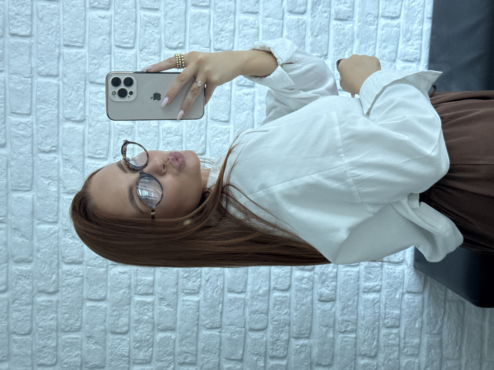 A young woman is captured in a selfie-style photograph, holding an iPhone and posing indoors. She is seen from the chest up, with long, straight brown hair and wearing a crisp white long-sleeved collared shirt. A section of a brown lower garment, possibly a skirt or pants, is visible.
The woman is taking the picture herself, holding a light-colored iPhone (likely a Pro model given the triple camera array) with her left hand, while her right hand's fingers are also visible near the device. The back of the phone prominently displays the Apple logo and the word "iPhone." She is wearing dark-rimmed glasses and has her lips playfully puckered, looking towards the phone's camera. Her right hand is adorned with multiple gold-colored rings on one finger and a single ring on another.
The background features a textured white brick wall, suggesting an interior setting in Chaikovsky, Russia. A darker, possibly black, element is visible on the far right, hinting at furniture or another wall section. The lighting appears soft and even, typical of an indoor environment.