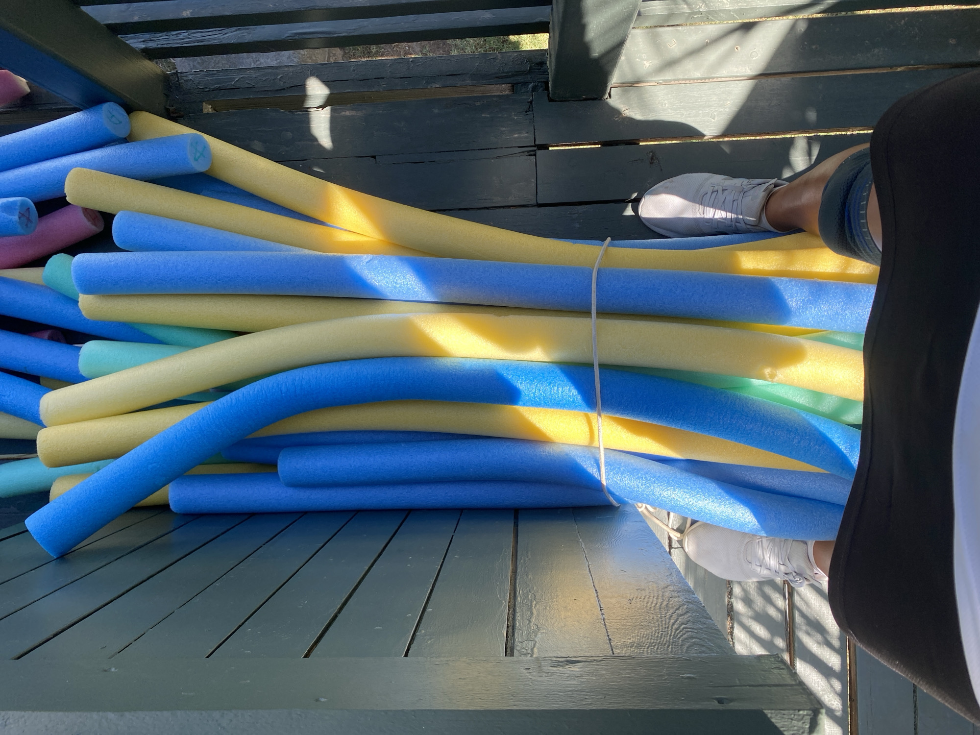 A bundle of blue and yellow pool noodles is tied together with a white string. They are leaning against a weathered, teal-painted wooden wall. The noodles are illuminated by dappled sunlight, casting shadows on the wooden planks beneath them. In the foreground, a person's legs are visible, wearing black shorts and white sneakers. The scene suggests an outdoor setting, possibly a porch or deck, with wooden railings to the right. The location is not Aksu, Turkey.