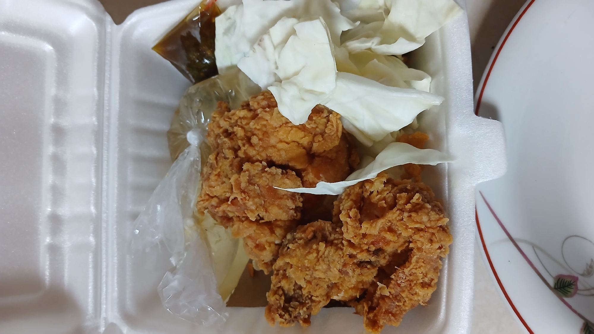 A polystyrene food container filled with fried chicken, pieces of white cabbage, and a small plastic bag of dark sauce is shown from an overhead perspective. The container also appears to contain a packet of yellow rice. The food is arranged inside the container, taking up most of the available space. The image provides no specific information about the location in Lhokseumawe, Indonesia, people, actions, or interactions.