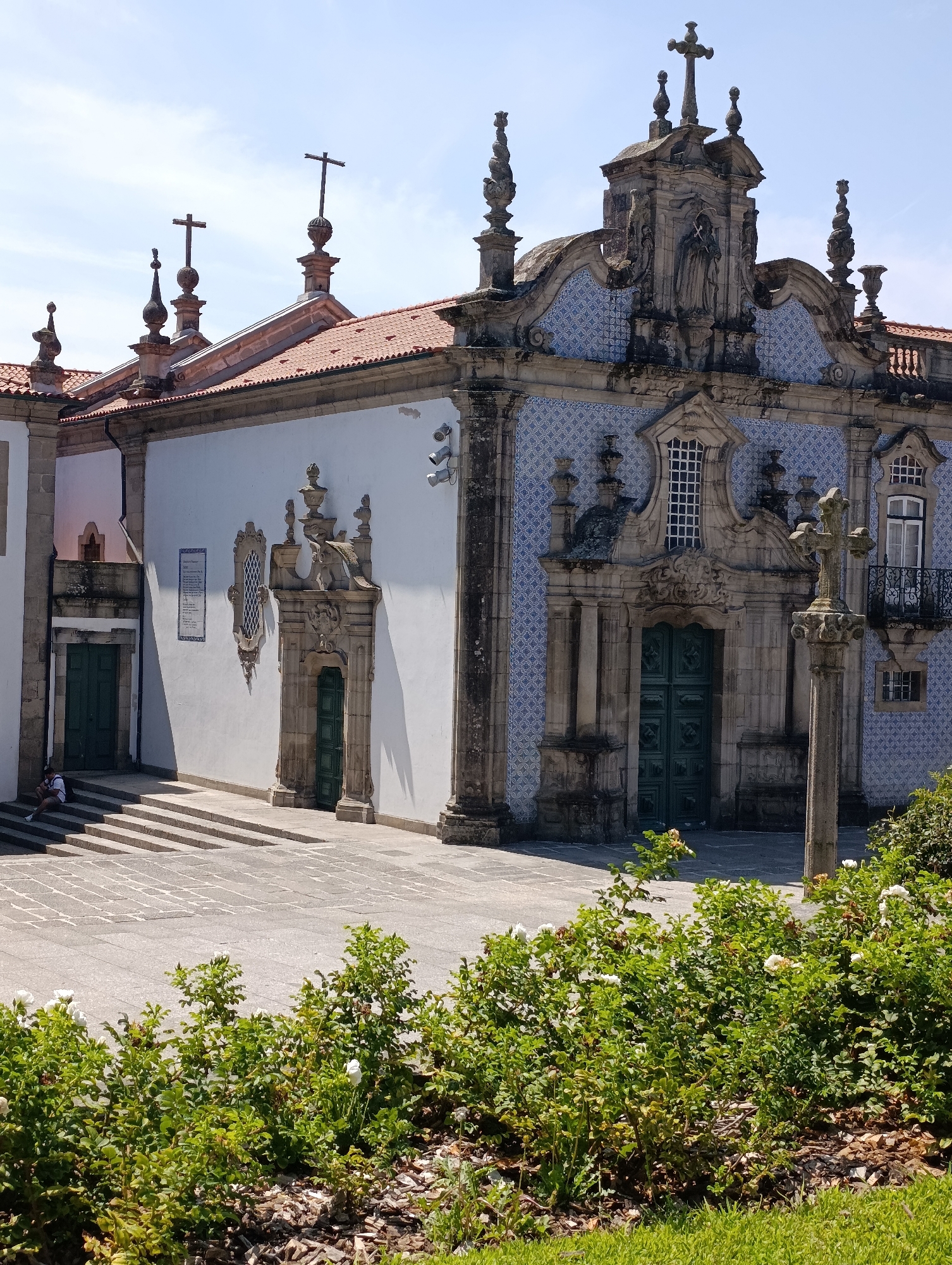 The image shows the exterior of the Santuário da Penha in Guimarães, Portugal. The sanctuary is a large, ornate building with a white facade, a red-tiled roof, and decorative blue tiles on its facade. A stone cross stands in front of the building, and a person is seated on the steps to the left. Lush green bushes and grass are in the foreground, and the sky is clear and blue. The architecture is Baroque, with intricate carvings and statues adorning the building. The overall scene suggests a peaceful, sunny day at a religious site.