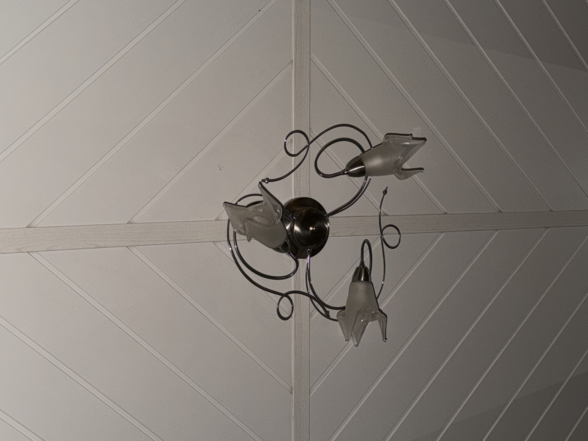 A three-arm ceiling light fixture is centrally positioned against a white wood-paneled ceiling. The fixture has a metallic silver finish and ornate, curved arms that extend outwards. Each arm holds a frosted glass lampshade shaped like a stylized flower or bud. The ceiling panels are arranged in a chevron pattern, meeting at a central ridge. The lighting appears to be dim, casting faint shadows. The scene depicts an interior shot looking directly upwards.