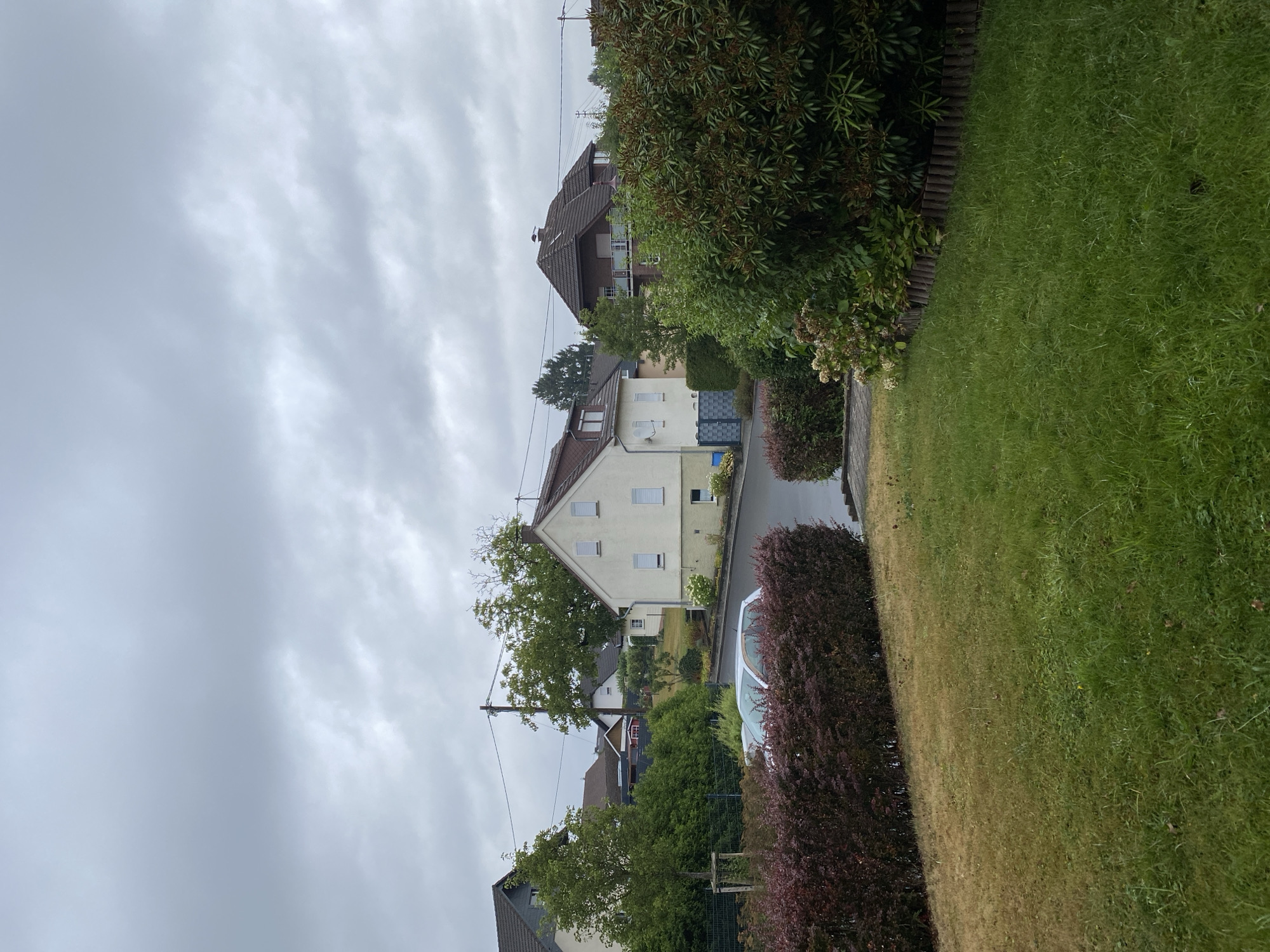 The image depicts a residential street in Flammersfeld, Germany, under an overcast sky. In the foreground, a well-maintained lawn transitions to a dark red hedge. Beyond the hedge, a light-colored car is parked on a wet asphalt road that curves uphill. Several houses are visible on either side of the road. A two-story house with a white facade and a brown roof is prominent on the right, featuring several windows with white shutters. To the left, another house with a grey roof and white walls is partially visible, with green trees and shrubs in front of it. The street is lined with trees and more houses can be seen further in the background, some with dark tiled roofs. Power lines are strung across the sky. The overall impression is of a quiet, suburban environment.