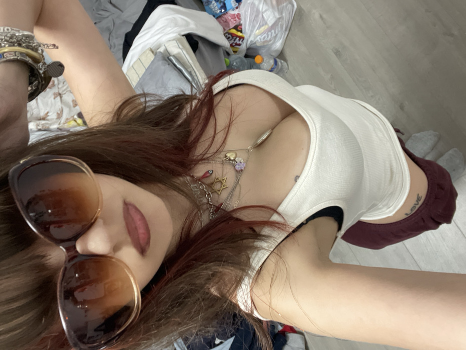 A woman with long, auburn-streaked hair lies at an angle, wearing large brown sunglasses. Her lips are painted a dark pink. She sports a white tank top with a low neckline, revealing necklaces and a tattoo on her upper chest. Her arm, adorned with bracelets, is raised. She has a "love" tattoo on her side. In the background, there are clothes, bags, and water bottles scattered on the floor. The floor itself appears to be made of a light-colored wood.