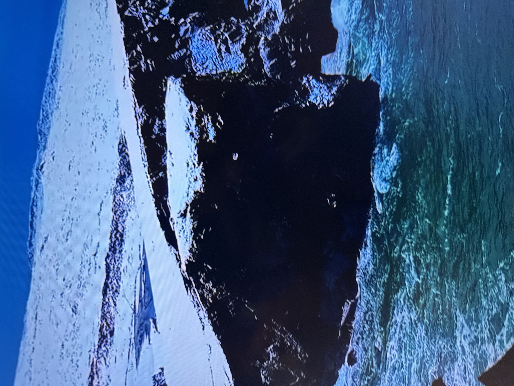 The image depicts a stark, cold landscape dominated by snow-covered terrain and dark, rocky cliffs meeting turbulent, greenish-blue ocean water. In the foreground, the rough sea churns with white foam, crashing against the base of a steep, dark cliff face. The cliff is partially covered in snow, with visible rocky textures and shadows. Beyond the cliff, a vast expanse of snow stretches inland, with undulating terrain and darker, possibly exposed rock formations or ridges breaking the white surface. The sky above is a clear, bright blue. The overall impression is one of a rugged, possibly Arctic or sub-Arctic coastal environment, likely a remote and undeveloped area. There are no visible signs of human presence, infrastructure, or any identifying features that would suggest a specific location within Almaty, Kazakhstan. Almaty is a landlocked city, situated in the foothills of the Tian Shan mountains, and is not characterized by coastal or oceanic landscapes.