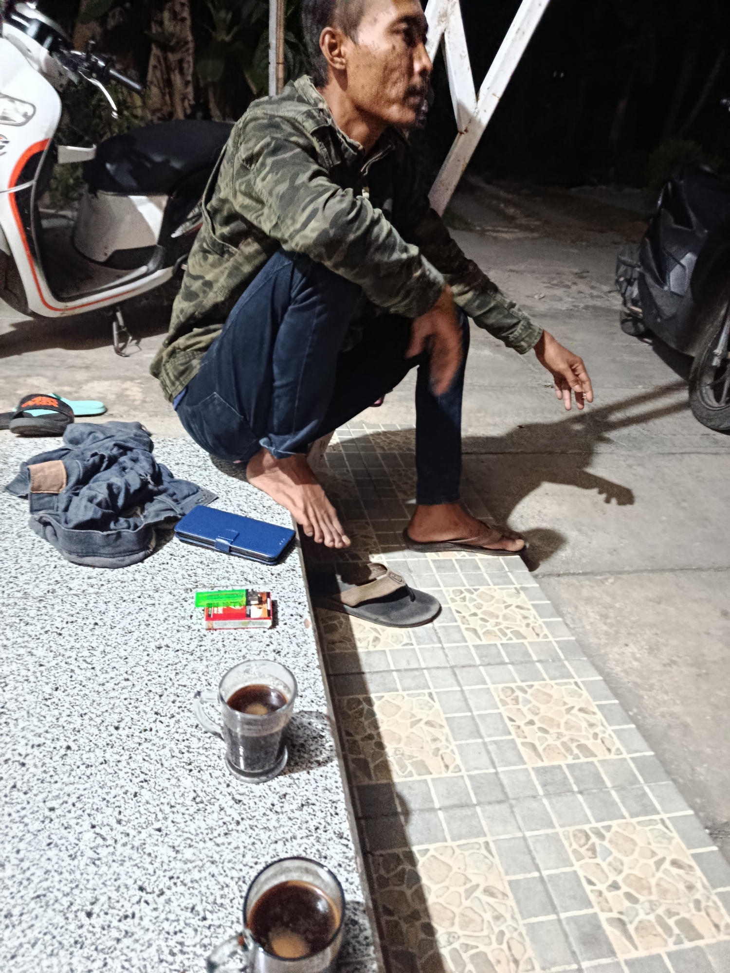 A man in a camouflage jacket and jeans is crouching on a textured surface, possibly a sidewalk or patio, in front of a tiled border. He is facing to the right, with his legs bent and one foot resting on a sandal. To his left, on the textured surface, is a pile of denim clothing, a blue smartphone case, and a packet of cigarettes next to a lighter. Two glasses of dark liquid, likely coffee, are also visible on the textured surface. In the background, a white scooter is parked to the left, and a motorcycle is partially visible to the right. The scene appears to be outdoors at night, with artificial lighting casting shadows. The location is identified as Sarwadadi, Indonesia.