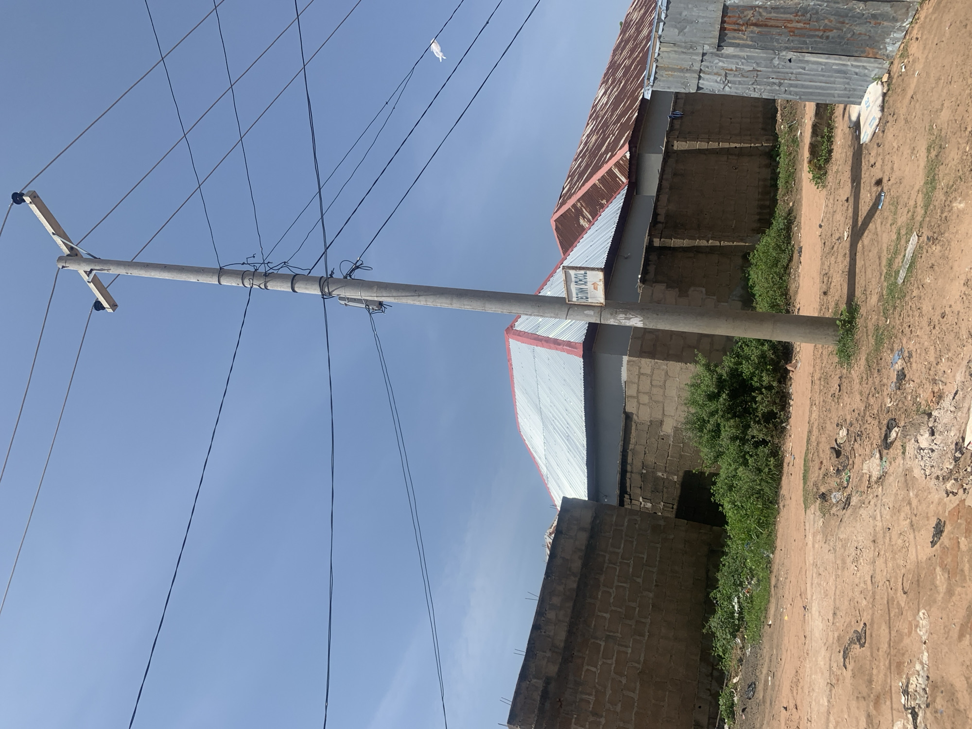 In Jauro Yinu, Nigeria, a scene unfolds beneath a clear blue sky, dominated by a concrete utility pole and overhead wires. The pole stands prominently, its top adorned with a wooden crossbar supporting insulators, with numerous black and lighter-colored wires radiating outwards. To the left of the pole, a low, brick wall made of light brown cinder blocks forms the boundary of a property. Beyond this wall, the roof of a building with a corrugated metal sheet and red trim is visible. A sign on the utility pole, visible in the middle right, reads "DESTINY SCHOOL" with an arrow pointing to the right. Further to the right, another building with a rusted, corrugated metal roof is partially visible. The ground in the foreground is a mix of reddish-brown dirt, scattered debris including plastic bags and other refuse, and patches of green grass and vegetation. To the far right, a structure constructed of corrugated metal panels stands as a freestanding unit. The overall environment suggests an outdoor, semi-urban or rural setting.