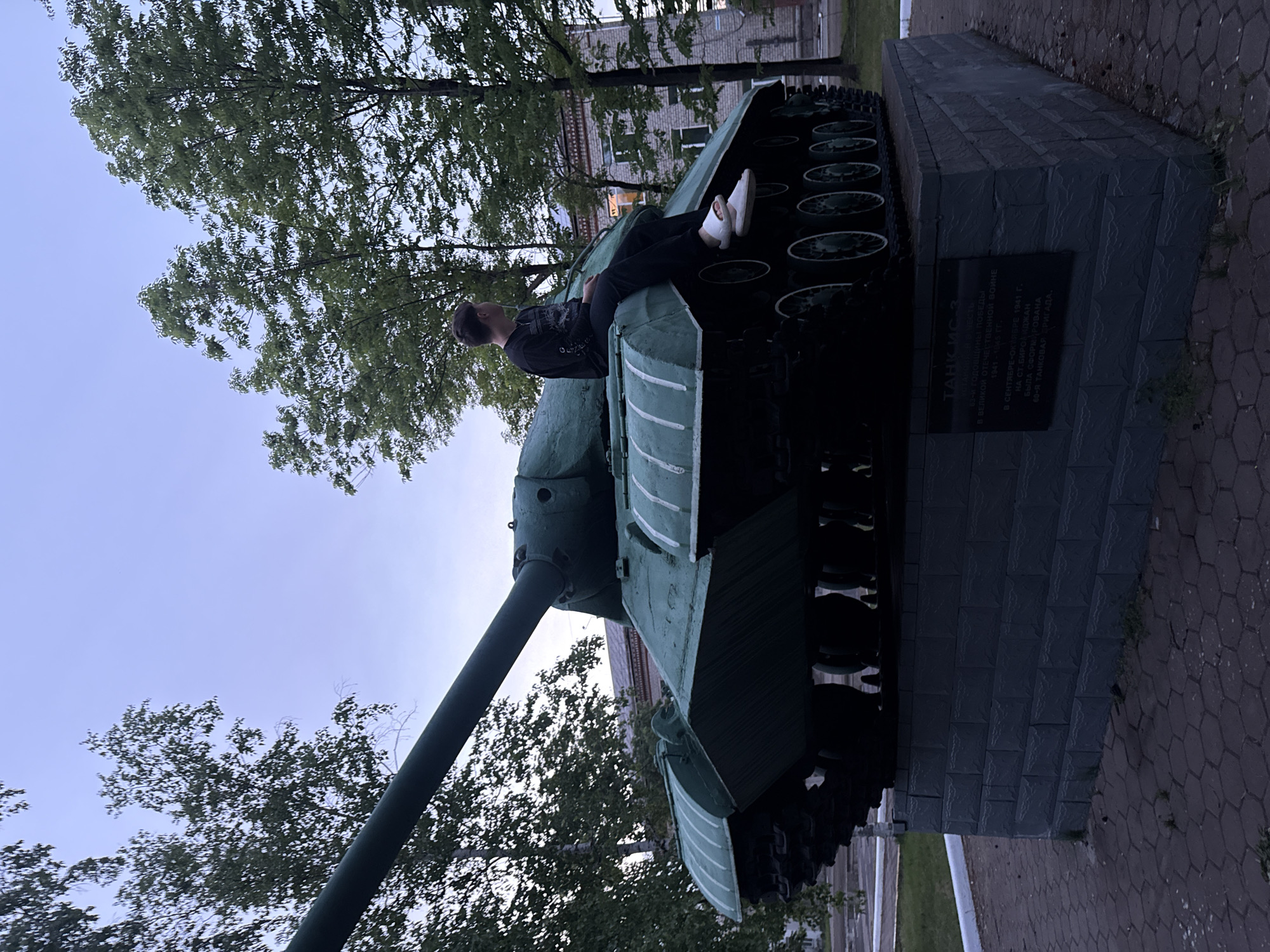 The image, taken in Birobidzhan, Russia, captures a young person sitting casually atop a green IS (Iosif Stalin) tank monument. The tank is painted a dark green with lighter green or white stripes on its side fenders, and its long gun barrel extends towards the left side of the frame. The person is dressed in dark pants and a dark long-sleeved shirt, wearing white slip-on shoes, and is looking up and to the left, towards the sky and trees.
The tank rests on a dark, stone-block pedestal. Surrounding the monument are numerous trees with dense green foliage, suggesting an outdoor park or public square. A multi-story building is visible in the background, partially obscured by the trees. The ground is paved with dark, possibly hexagonal, pavers.
The lighting indicates a soft, diffused light, characteristic of an overcast day or late afternoon/early evening.
A black plaque is affixed to the tank's pedestal. The text on the plaque, in Cyrillic, translates to: "IS TANK. In honor of the 63rd anniversary of victory in the Great Patriotic War 1941-1945. In September-October 1941, at Birobidzhan Station, the 60th Tank Brigade was formed." This plaque identifies the tank as a memorial dedicated to a specific historical event involving the 60th Tank Brigade in Birobidzhan during World War II. The person appears relaxed while interacting with this historical monument.