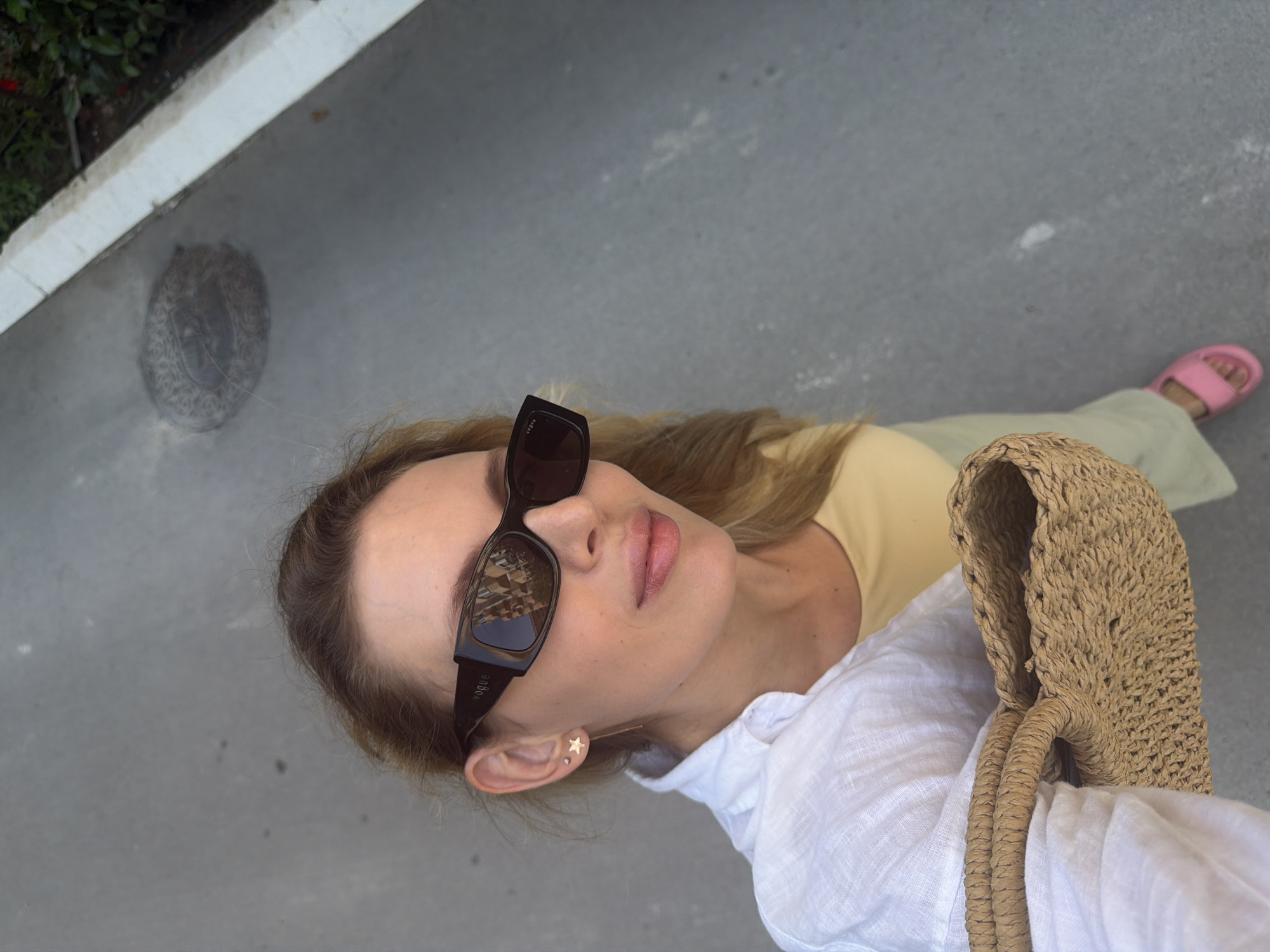In Budva, Montenegro, a woman with blonde hair, wearing sunglasses, a white shirt, and a yellow top, holds a straw bag. She is wearing pink slides and a star-shaped earring. The background shows a street with a manhole cover and greenery.
