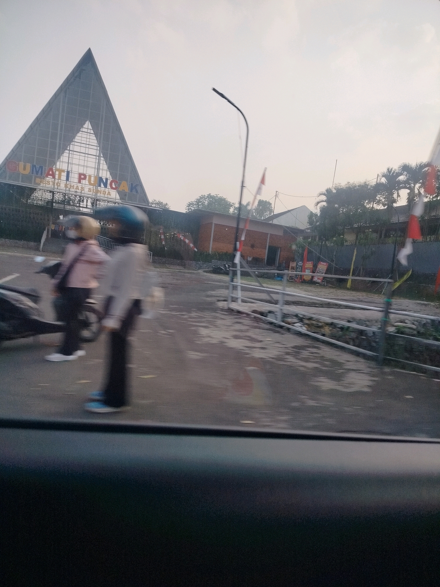 The image depicts the entrance to Gumati Puncak, a tourist destination in Megamendung, Indonesia. A large, triangular architectural structure with the words "GUMATI PUNCAK" emblazoned in colorful letters dominates the background. Below it, smaller text reads "DESTO KHAS SUNDA". In the foreground, two individuals on a scooter are visible, one wearing a light-colored jacket and a yellow helmet, the other in a light-colored shirt and a dark blue helmet. They appear to be stationary. To the right, a street lamp and several Indonesian flags are present. The environment appears to be overcast, with trees and buildings visible in the distance. The road surface is wet, suggesting recent rain. The image is taken from a slightly elevated, moving perspective, likely from inside a vehicle.