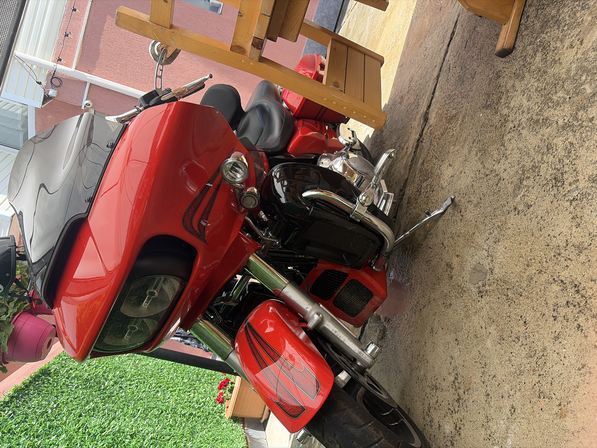 A bright red touring-style motorcycle is parked on a textured concrete surface during daylight hours in Tsarevo, Bulgaria. The motorcycle features a large, integrated front fairing with a black upper section and a dark tinted windshield. Dual rectangular headlights are recessed into the front fairing. The red bodywork, including the front fairing and fender, is adorned with black pinstriping and custom flame-like graphics. Chrome components are visible on the front forks, engine casing, foot controls, and exhaust system. A black two-up seat is present. The motorcycle is supported by its kickstand, which is deployed on the left side.
To the immediate left of the motorcycle, a vertical surface is covered with a vibrant green artificial hedge. Above this hedge, a pink plastic pot containing a plant is suspended. At the base of the hedge, a terracotta pot holds red blooming flowers. To the right of the motorcycle, a light-colored wooden structure, possibly part of outdoor furniture, is partially visible.
The background consists of a building with light pink/peach colored stucco walls. Attached to the wall are a white downspout or support column, a small white outdoor security camera, and a string of outdoor lights. A white-framed window is visible in the upper section of the building wall. The ground beneath the motorcycle and foreground objects appears to be stained or wet in certain areas. No people are present in the scene.