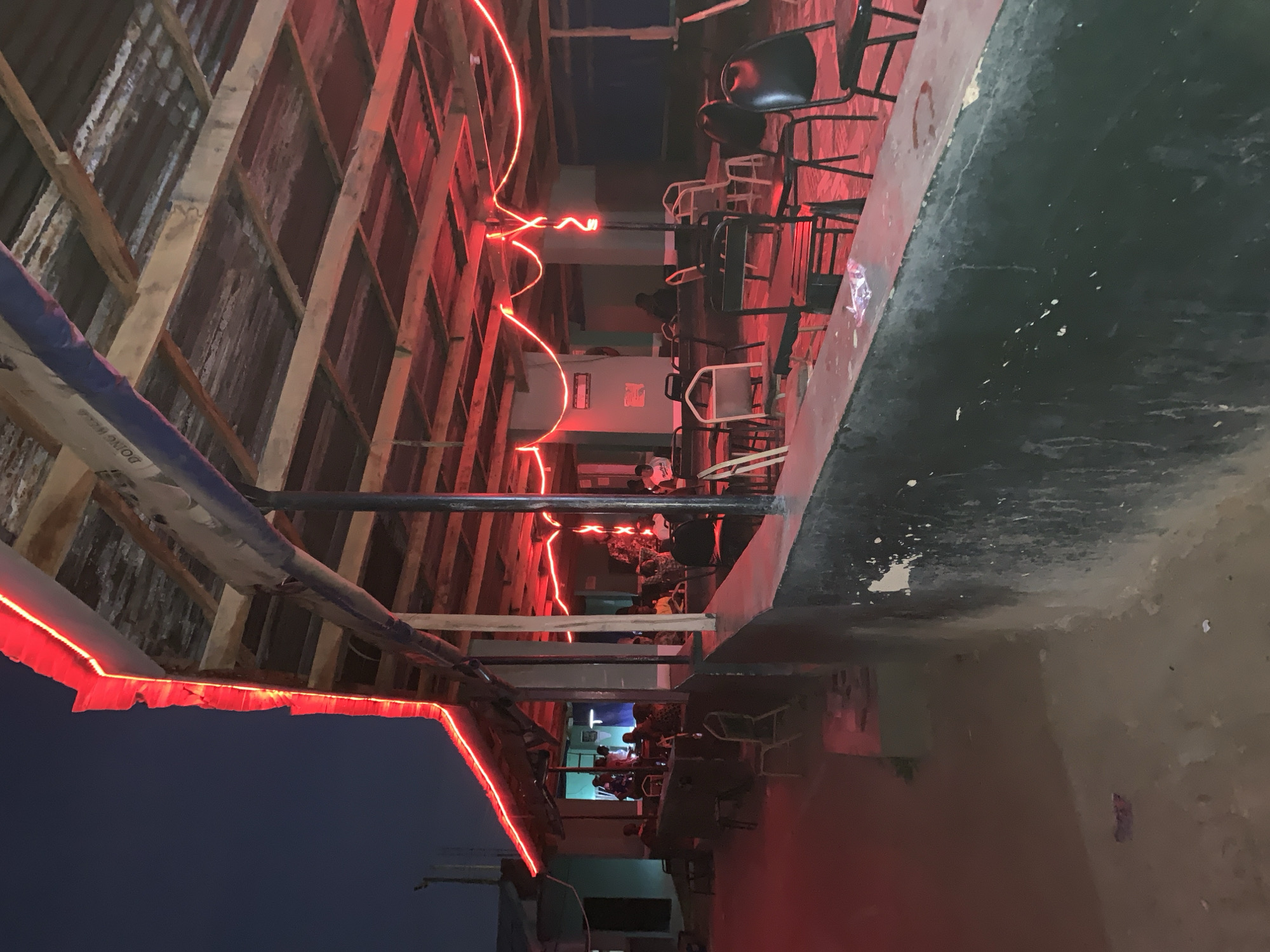 The image shows an open-air establishment in New Stadium, Nigeria, likely a bar or restaurant, at night. The space is partially enclosed with a corrugated metal roof supported by wooden beams. Red neon lights run along the edges of the roof. Tables and chairs are visible, with people seated at some of them. The image is taken from a skewed angle, looking down the length of the space. The night sky is visible in the lower left corner.