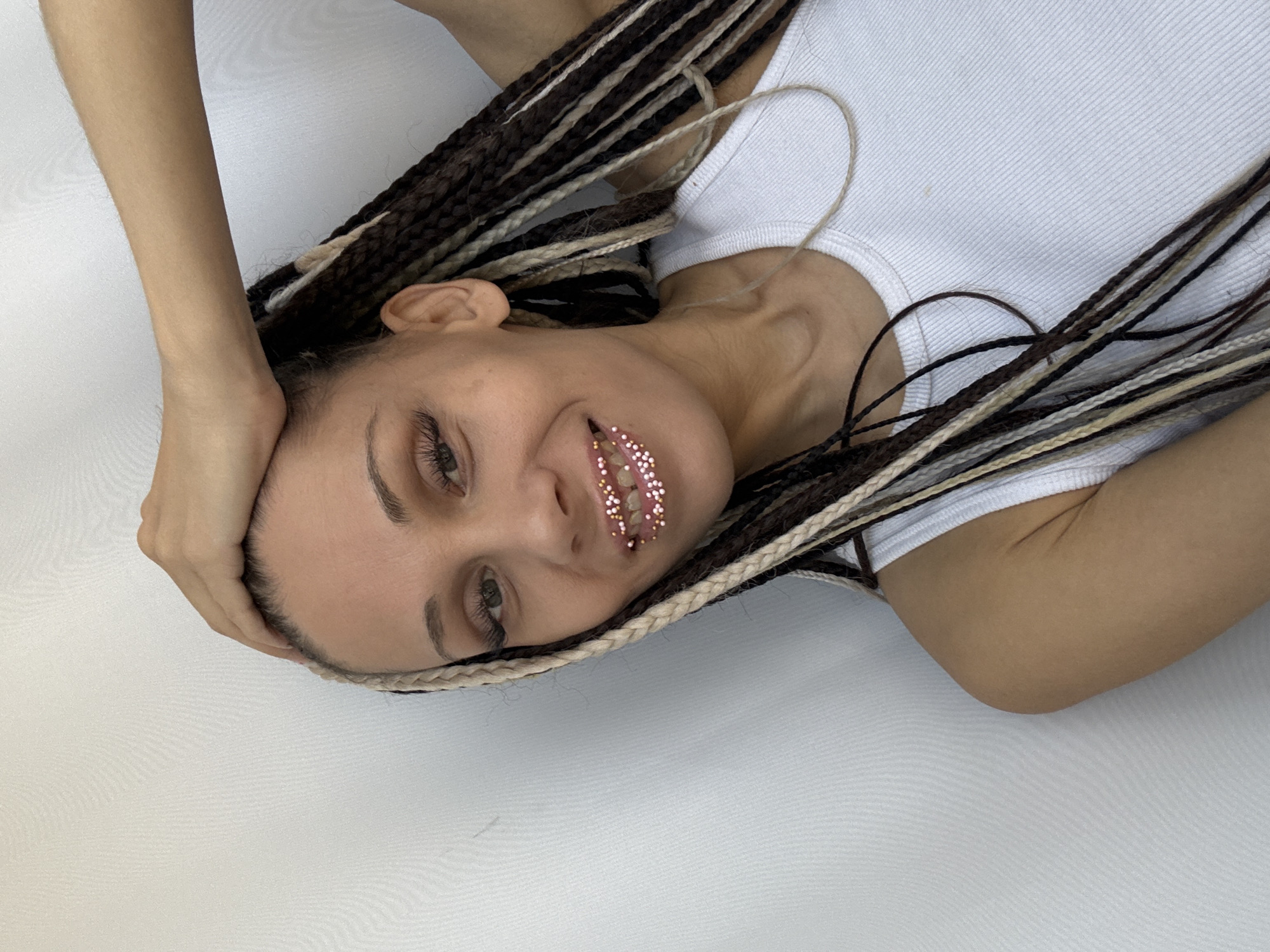 A woman with braided hair, some strands blonde and some brown, is lying on a white surface. She is wearing a white tank top. Her left hand is resting on her forehead. There are small, pink decorations on her lips. The location is unknown.