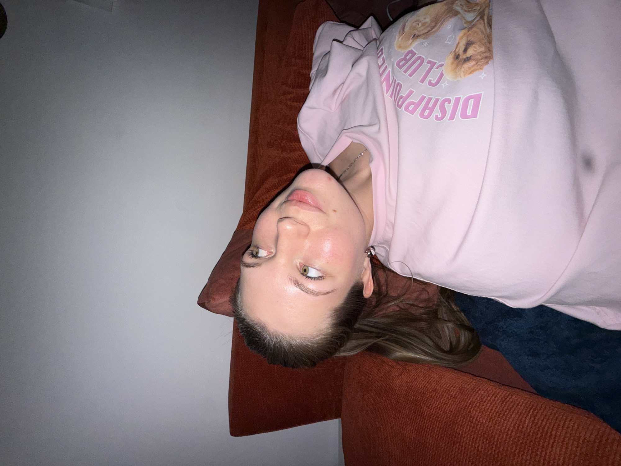 An indoor scene within Vozdovac Urban Municipality, Serbia, depicts a female individual positioned against an orange-red upholstered sofa. Her head is tilted to the left from the viewer's perspective, with her face oriented upwards and to the right, exhibiting a slight smile. She possesses light skin, light-colored eyes, and dark brown hair pulled back. She is wearing a light pink long-sleeved shirt with "DISAPPOINTMENT CLUB" printed in pink text and an image of two brown rabbit-like figures on the chest. A thin silver-colored chain necklace is visible around her neck, and a silver hoop earring is present in her visible right ear. Dark blue fabric, likely pants, is visible below her waist. The background behind the sofa is a plain, light grey wall. The overall lighting is dim.