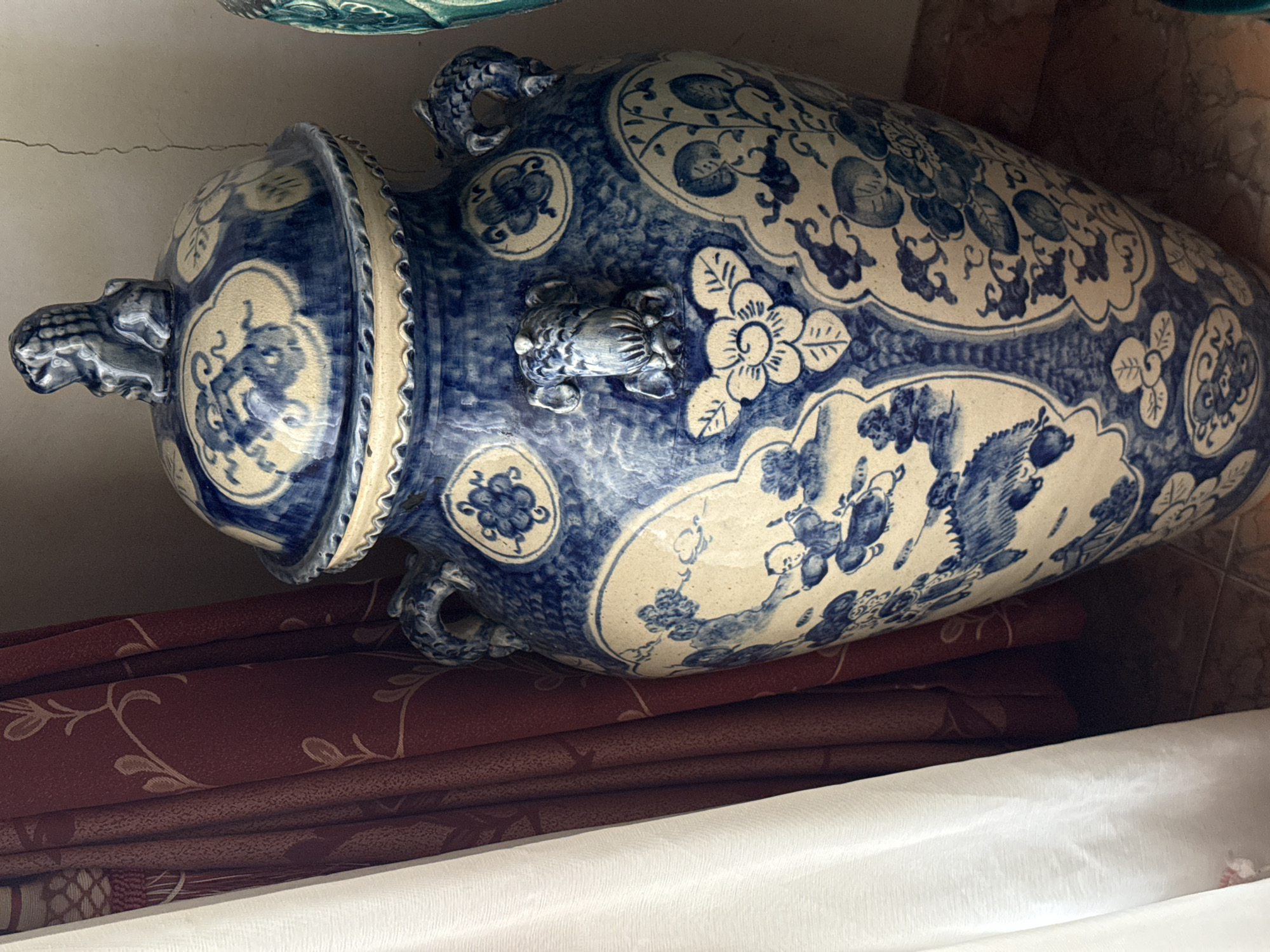 A close-up, slightly angled, medium shot captures a large, antique, blue and white porcelain vase with a lid. The vase is ornately decorated with a faded floral pattern and what appear to be figures in a landscape. Two foo dog finials are attached to the lid and the body of the vase, respectively. The vase rests on a tiled floor, partially obscured by a white sheer curtain to the left and a deep red curtain with a faint floral pattern to the right. The background is a textured, light-colored wall. The scene is static and indoors, likely in a domestic setting in Bireuenmeunasahblang, Indonesia.