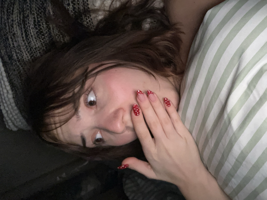 A woman with brown hair and bangs lies on her side, her head resting on a striped pillow. She gazes at the viewer with wide eyes, her hand raised to her cheek. She has red nails decorated with white polka dots. The background includes a textured gray blanket.