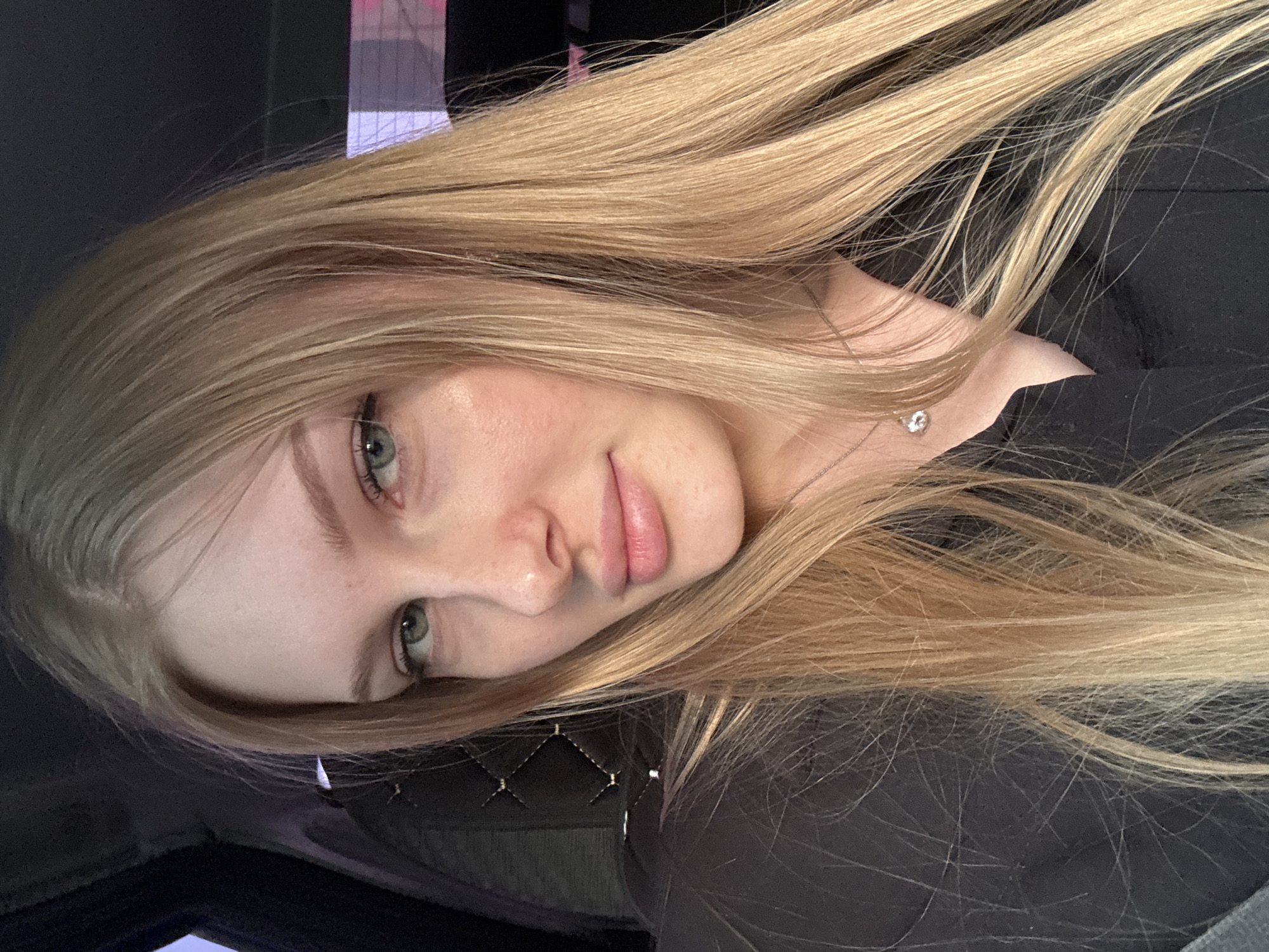The image is a portrait of a fair-skinned woman with long, light blonde hair. She has light-colored eyes and is wearing a necklace with a small pendant. The woman is wearing a black top. The background appears to be a vehicle interior, with black upholstery featuring a quilted pattern visible behind her. The setting is possibly in Perm, Russia.