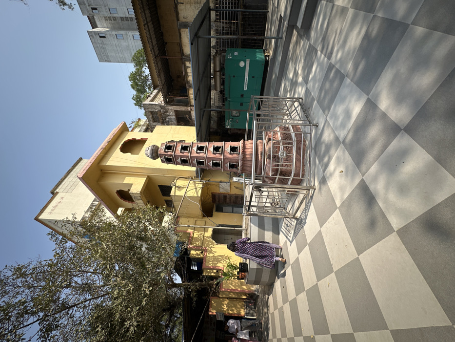 A unique, tiered tower with ornate carvings sits enclosed by a silver cage on a checkered gray and white tile surface. Beside the tower, a yellow building with arched windows rises behind a bare tree. In the background, a green generator stands near an older, dilapidated building. A woman in a purple and white outfit approaches the tower. The sky is clear and blue.