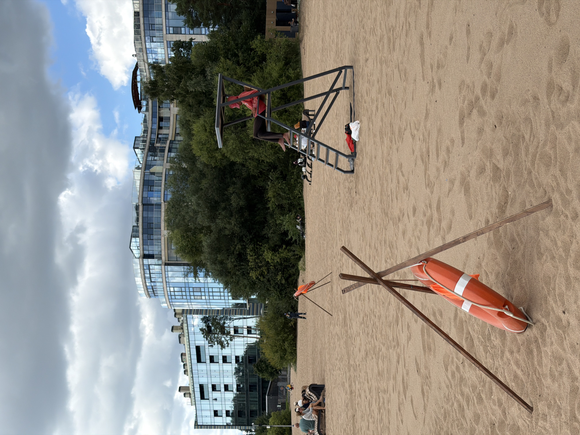 The image depicts a sandy beach scene in Repino, Russia, with modern, glass-fronted buildings in the background. In the foreground, an orange life preserver with white stripes is propped up by three wooden stakes. Further back, a lifeguard sits in a raised observation tower, wearing a red top and a red cap. Several people are visible on the beach, some sitting on benches and others scattered around. Trees and vegetation border the beach behind the people and lifeguard tower. The sky is partly cloudy.