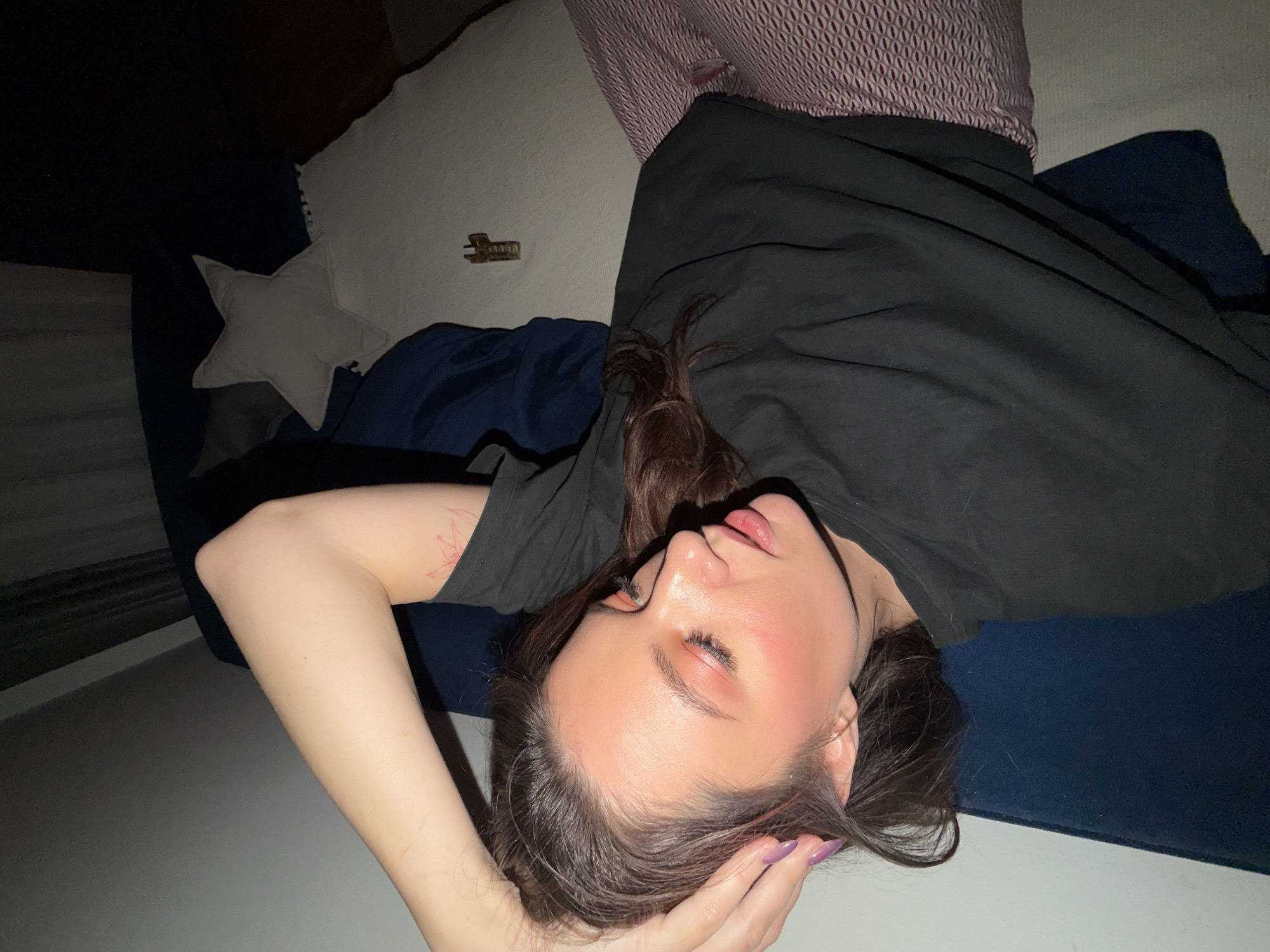 A woman is lying on a sofa. Her arm is bent above her head, and her head is tilted back. She is wearing a black t-shirt. The sofa has several pillows. The location cannot be determined.
