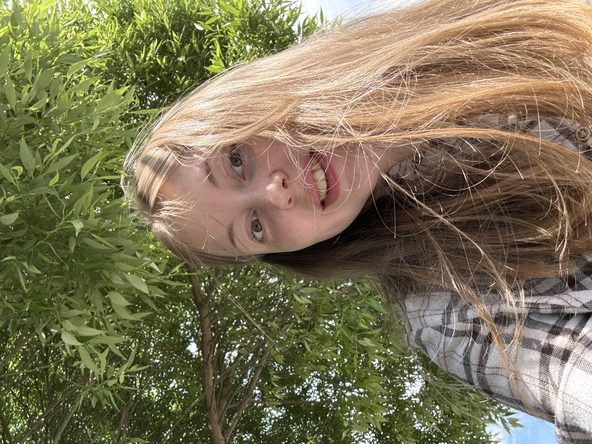 A young woman with long, light brown hair and fair skin with freckles is captured from a low angle, looking upwards. She is wearing a plaid shirt. Her mouth is slightly open, showing her teeth, and she appears to be sticking her tongue out slightly. Her eyes are green. She is outdoors, surrounded by lush green leaves of a tree overhead, with a glimpse of a blue sky. The scene suggests a casual, possibly sunny day. The location is unknown.