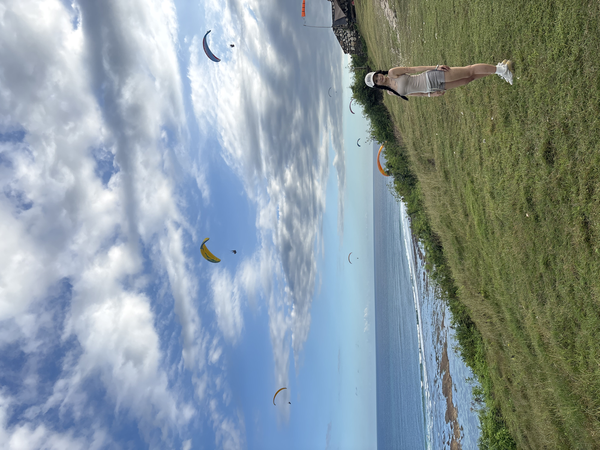 The photograph depicts a woman standing on a grassy cliff overlooking the ocean in Kuta Selatan, Indonesia. She is wearing a white baseball cap, light-colored tank top, shorts, and sneakers. Several paragliders are visible in the sky above the ocean. The sky contains cumulus clouds. The water has some waves breaking on the shoreline. The grass extends back, forming a small cliff where the woman is standing. The image is rotated so that the sky is to the left and the grass is to the right.