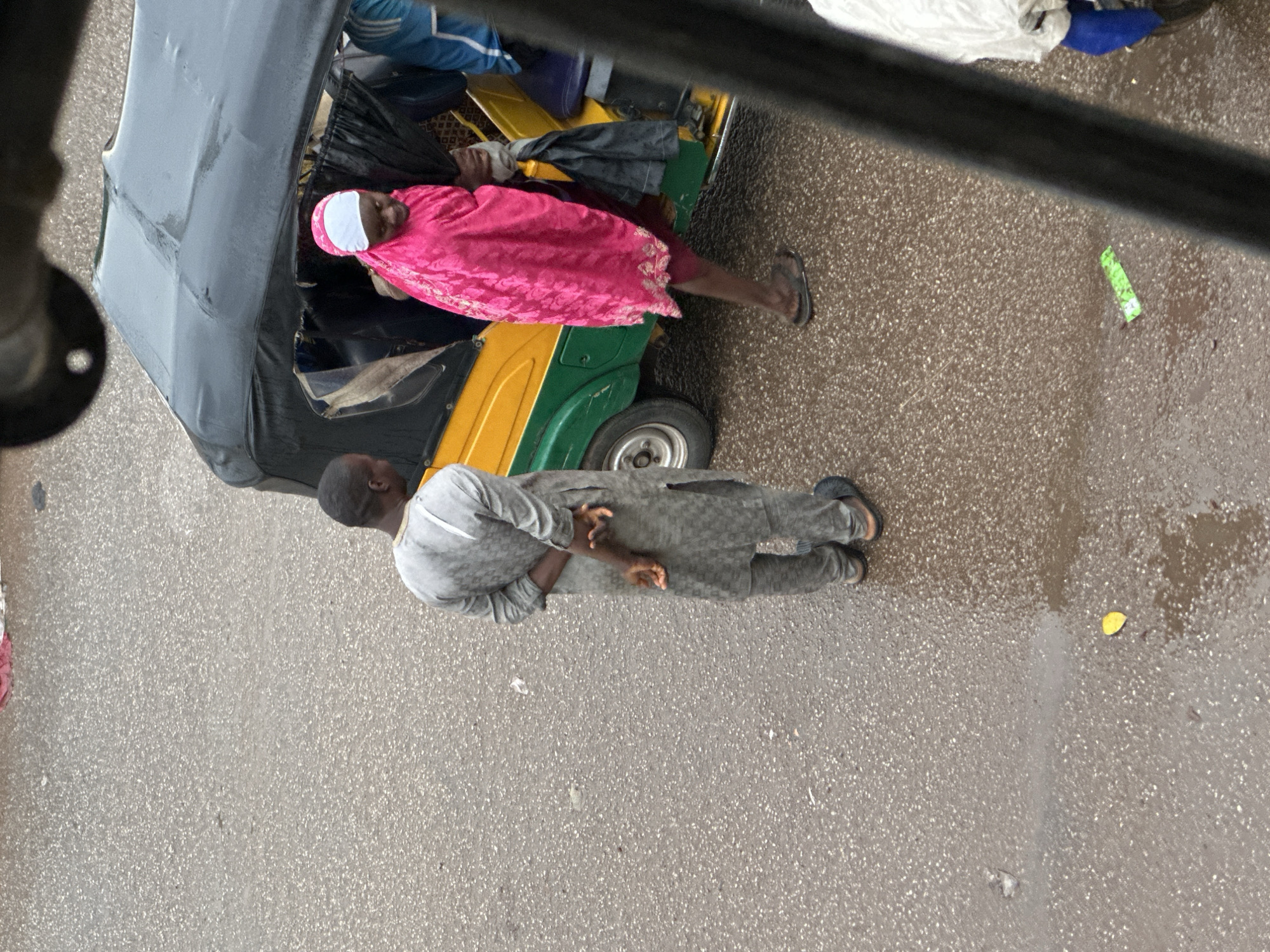 In Bukuru, Nigeria, a man in a grey patterned tunic and trousers stands with his hands behind his back, facing away from a three-wheeled vehicle. A woman, dressed in a bright pink hijab and matching wrap, is partially obscured by the vehicle's roof as she appears to be boarding or disembarking. The vehicle is yellow and green, with a black canopy. The ground is wet and appears to be asphalt, with a yellow leaf visible on the surface. The overall lighting suggests an overcast day.