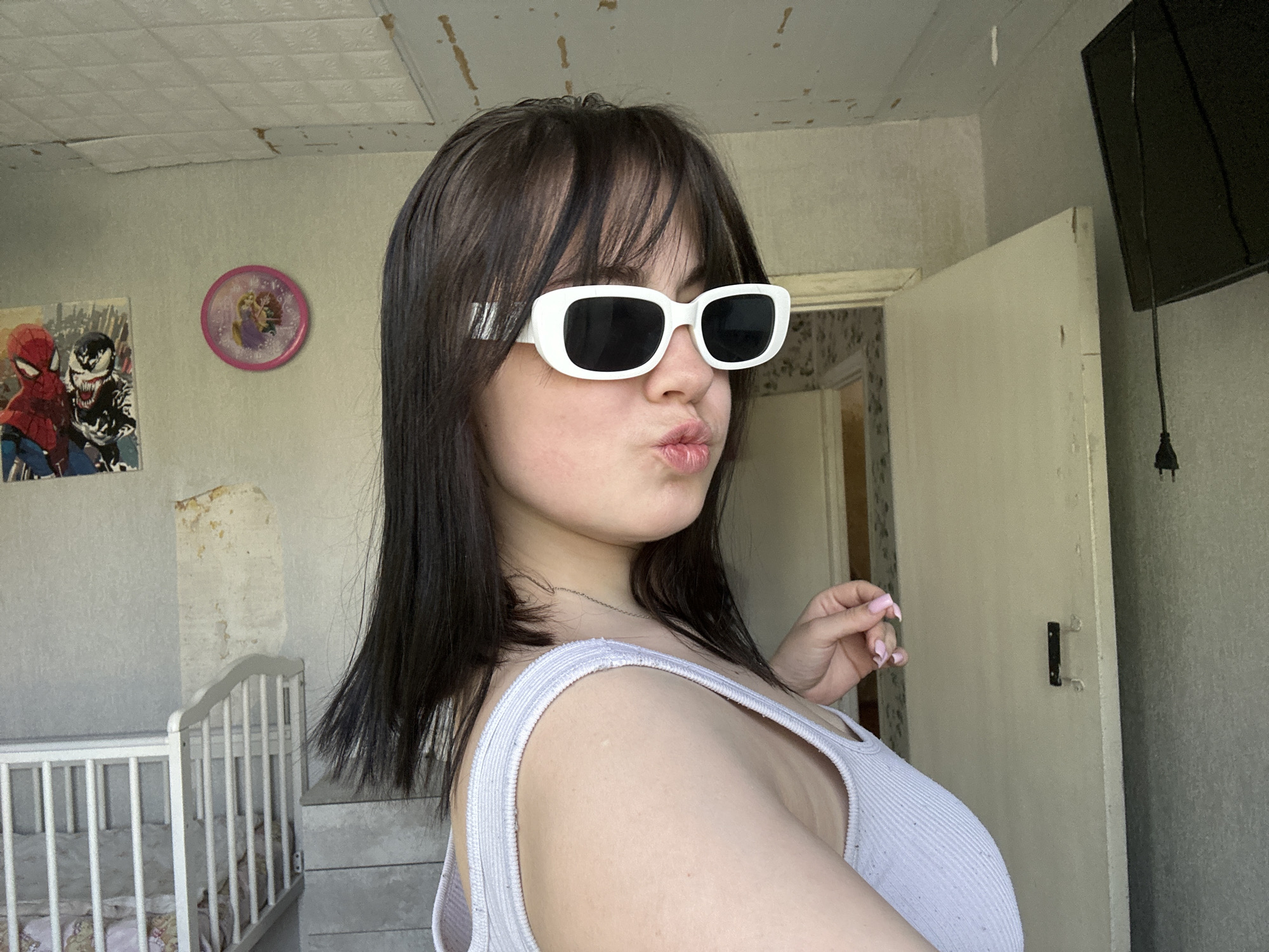 A young woman wearing white-rimmed sunglasses and a light-colored tank top is posing for a photo. She is pursing her lips in a kiss. Behind her is a room with a crib, a peeling wall with a Spider-Man poster and a Rapunzel clock. There is a television hanging on the wall and an open doorway. The location is likely a residential interior, based on the furniture and decor. The city of Chaikovsky, Russia is not visually confirmed.