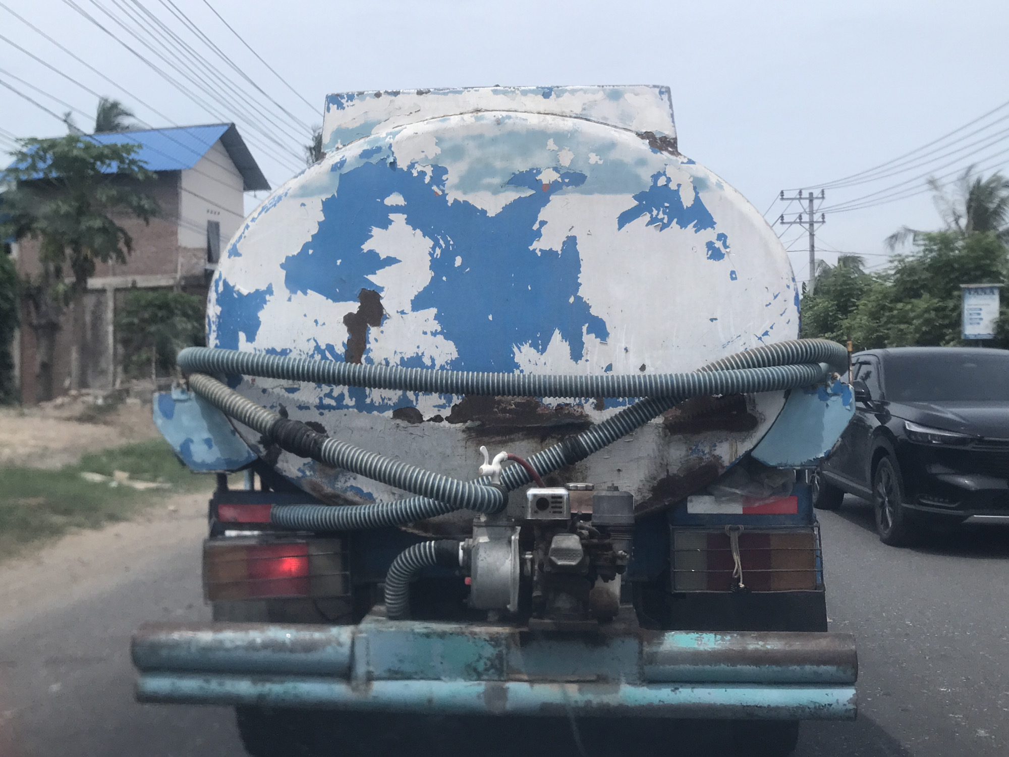 A close-up, rear-facing view of a water truck traveling down a road in Lhokseumawe, Indonesia. The truck features a large, cylindrical tank with peeling white and blue paint. A series of corrugated hoses run across the back of the tank, connecting to a small engine and pump mounted on the truck's bed. The truck's rear bumper is light blue and shows signs of rust. One taillight is illuminated. A dark-colored SUV is visible in the background, along with buildings, trees, and utility poles. The sky is overcast.