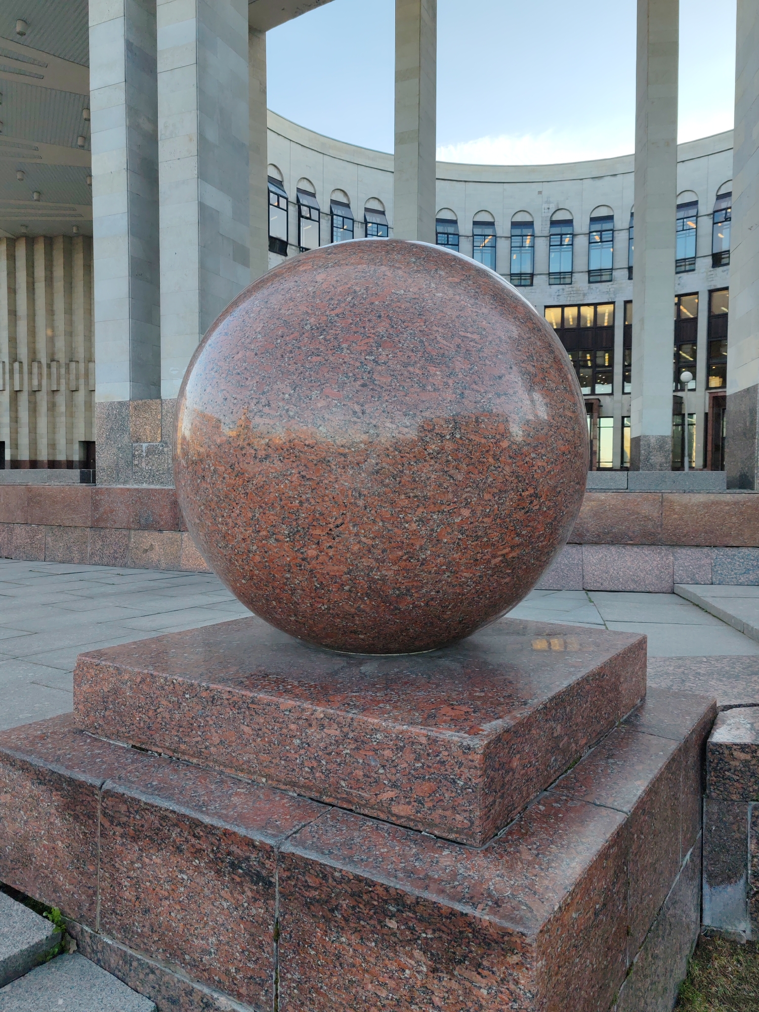 The image depicts a large, polished granite sphere atop a multi-tiered granite base, located in Saint Petersburg, Russia. The sphere is dark red with black speckles. The base is constructed from the same material, consisting of stacked rectangular slabs. The backdrop features a modern architectural structure with tall, white columns and a curved facade with windows. The ground is paved with grey stone slabs. The sky is visible in the background. There are no people visible in the image.