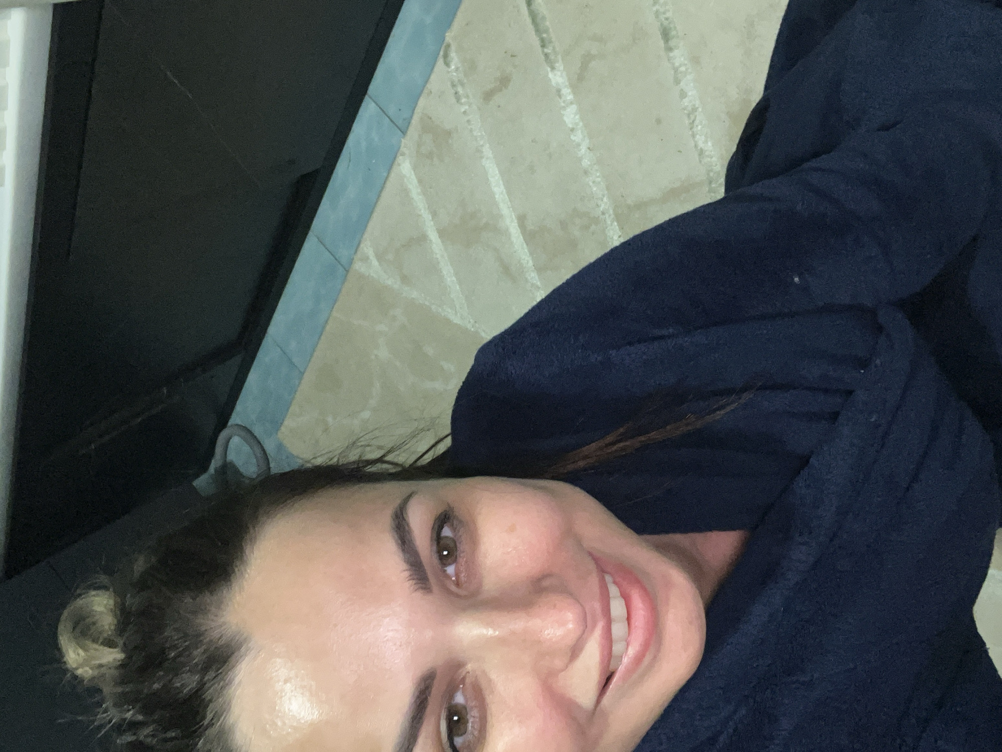 A close-up, high-angle shot shows a woman with light brown eyes and dark brown hair pulled into a messy bun. She is smiling and wearing a dark blue robe. The background consists of a tiled floor with a light brown hue and a darker, possibly black, surface above. The lighting is overhead and bright, creating a slight shine on the woman's face.