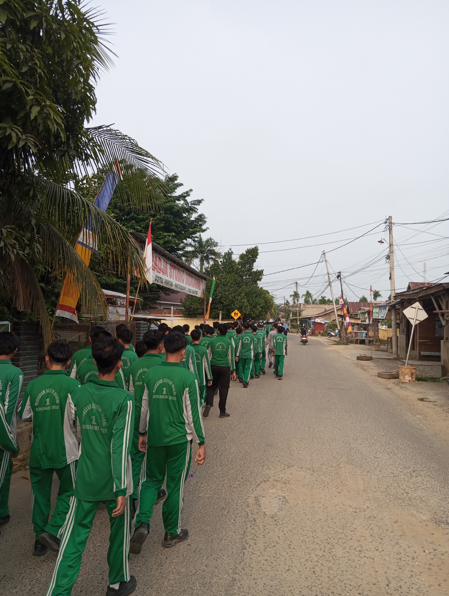 A group of students wearing matching green tracksuits with white stripes are walking down a paved road in a town, likely Pematang Reba, Indonesia. The students, predominantly male, are walking in formation from left to right of the frame, their backs to the camera. The tracksuits have text on the back. The road is lined with trees on the left, including palm trees and a large leafy tree, and buildings and power lines on the right. The sky is overcast and bright white. In the background, there are more trees, houses with corrugated metal roofs, and a few motorcycles. A banner with Indonesian text and flags are visible on the left side of the road. There are street signs and utility poles along the road. The environment appears to be a rural or semi-urban area.