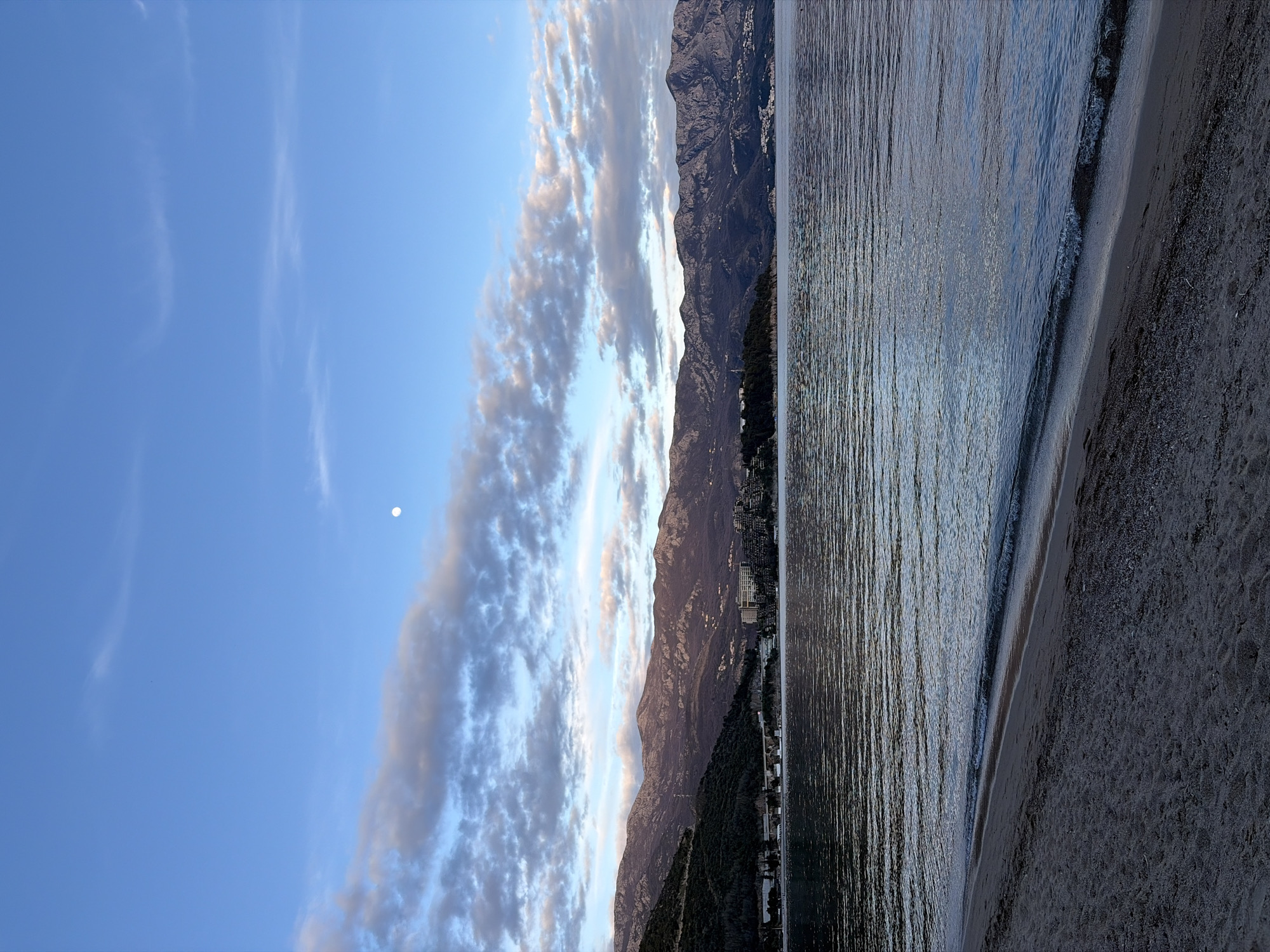 The image shows a view of Budva, Montenegro, at sunset or sunrise. The scene includes the sea, a beach with dark sand, and mountains in the background. The sky has a mix of blue and white clouds with a hint of pink or orange from the sun. A small white dot, likely the moon, is visible in the sky. The buildings of Budva are nestled between the mountains and the sea. The water is calm with small waves reaching the shore.