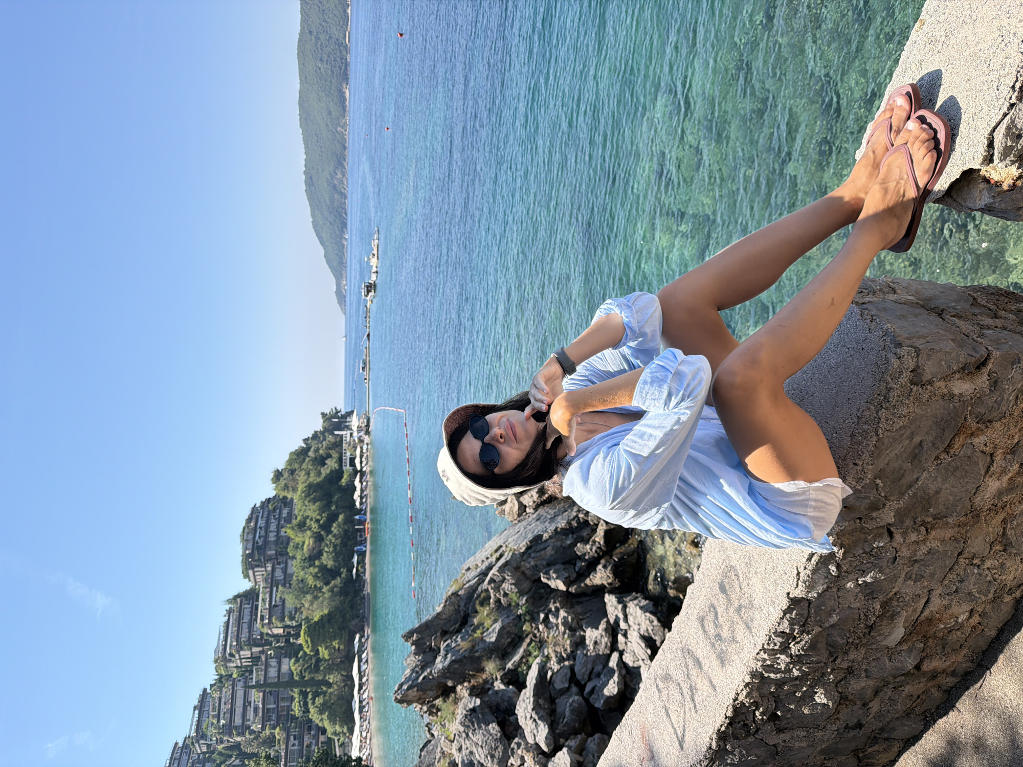 A woman is sitting on a stone wall overlooking the sea in Boreti, Montenegro. She is wearing a light blue oversized shirt, white shorts, a bucket hat, and sunglasses. The sea is a vibrant turquoise color, and there are buildings built into the hillside in the background. The woman is looking at the camera and smiling. The overall scene is peaceful and idyllic.