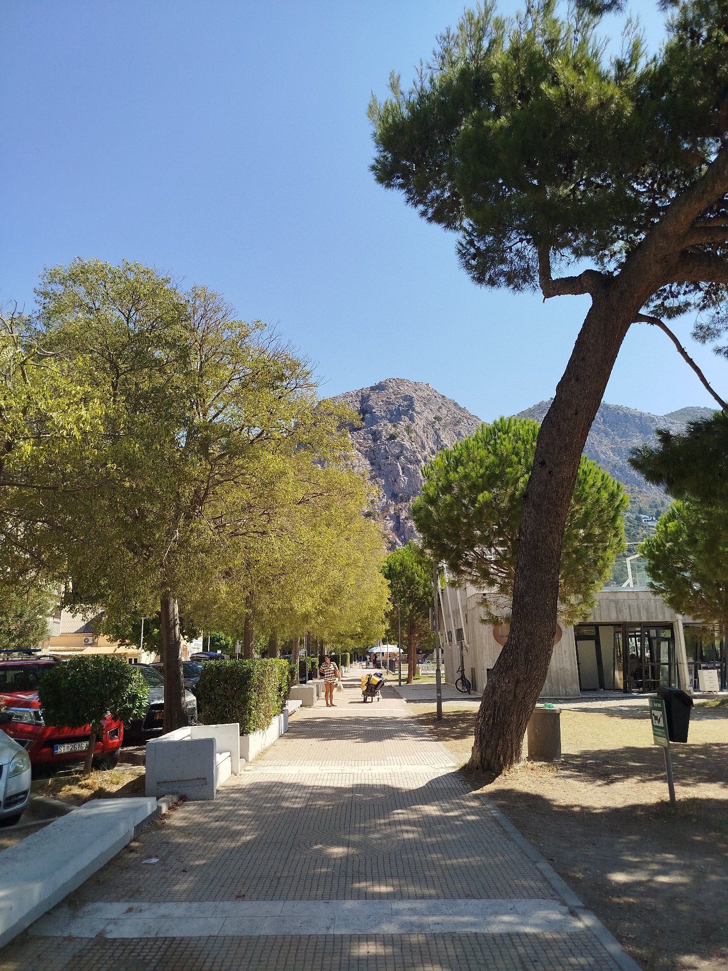 The image depicts a street scene in Omiš, Croatia, on a sunny day. A paved pedestrian walkway lined with trees runs parallel to a road. On the left side of the walkway, cars are parked, and there are some low hedges. A red car with license plate ST 2616 A is visible. On the right side of the walkway, there is a concrete bench, a trash can, and a building with large windows. In the background, there are mountains. A person is walking on the walkway, and a person is pushing a yellow cart. The sky is clear blue.