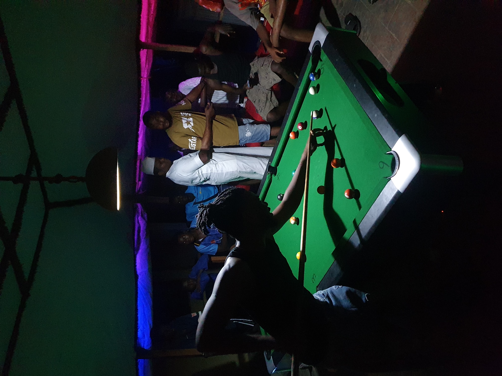This media file captures an indoor night scene in Lokoja, Nigeria, depicting a social gathering around a pool table. The main subject is a person, likely male, in the foreground on the right, silhouetted against the bright green pool table as they line up a shot with a cue stick. Several pool balls are visible on the table, including white, blue, orange, and striped balls.
In the mid-ground and background, a group of approximately eight to ten other individuals, mostly men, are gathered. Some are watching the game, while others appear to be socializing. Notable figures include a man in a yellow/khaki t-shirt with the text "EASTAR" printed on it, and another man wearing a white traditional garment and a white cap. The atmosphere appears casual and relaxed.
The setting is dimly lit by artificial light, with prominent colored lighting creating a vibrant mood. Pink and purple lights illuminate a wall section on the right, while blue and green hues are visible on the left, suggesting a bar, club, or recreation center. The walls appear to be roughly textured or unfinished. A metal light fixture hangs from the ceiling on the left side of the frame. The time of day is clearly evening or night, given the reliance on artificial lighting.