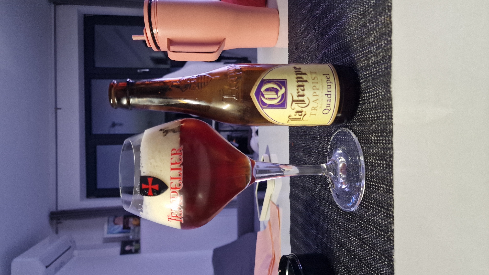 The image presents a tabletop scene. A half-full glass of beer, branded "Tripel Le Leflier," stands alongside a bottle of "La Trappe Trappist Quadrupel" beer. A pink object sits above the bottle. The tabletop has a dark, textured surface. The background includes a window, an air conditioning unit, and framed pictures, indicating an indoor setting. There's no explicit information to confirm the location in the city of Sofia, Bulgaria. No people or interactions are visible in the image.