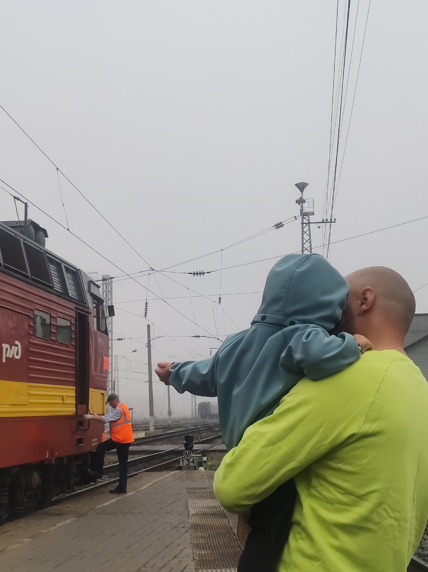 In Kirov, Russia, on a foggy day at a train station, a man holds a child in his arms. The child, wearing a light blue hooded sweatshirt, points towards a red and yellow train. A man in an orange safety vest and dark pants is visible near the train, appearing to be stepping onto it. The train is on tracks, and overhead power lines are visible against the hazy sky. The platform is paved with brick. The scene suggests a moment of observation or a departure.