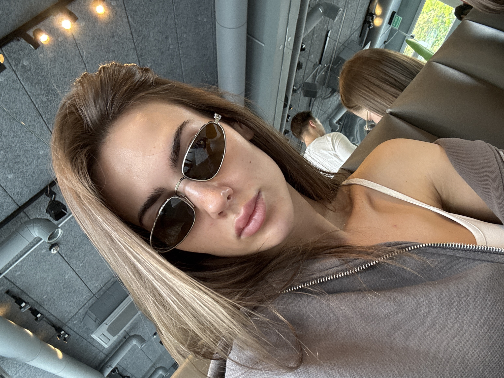 The image is a selfie of a fair-skinned young woman with light brown hair, wearing sunglasses and a grey zip-up top. She has a nose ring. In the background, there are two other people, one with brown hair and the other with their back to the camera wearing a white shirt. The location appears to be a cafe or restaurant in Warsaw, Poland, given the presence of modern industrial ceiling fixtures, ductwork, and the possible reflection of a Warsaw park in the mirror.