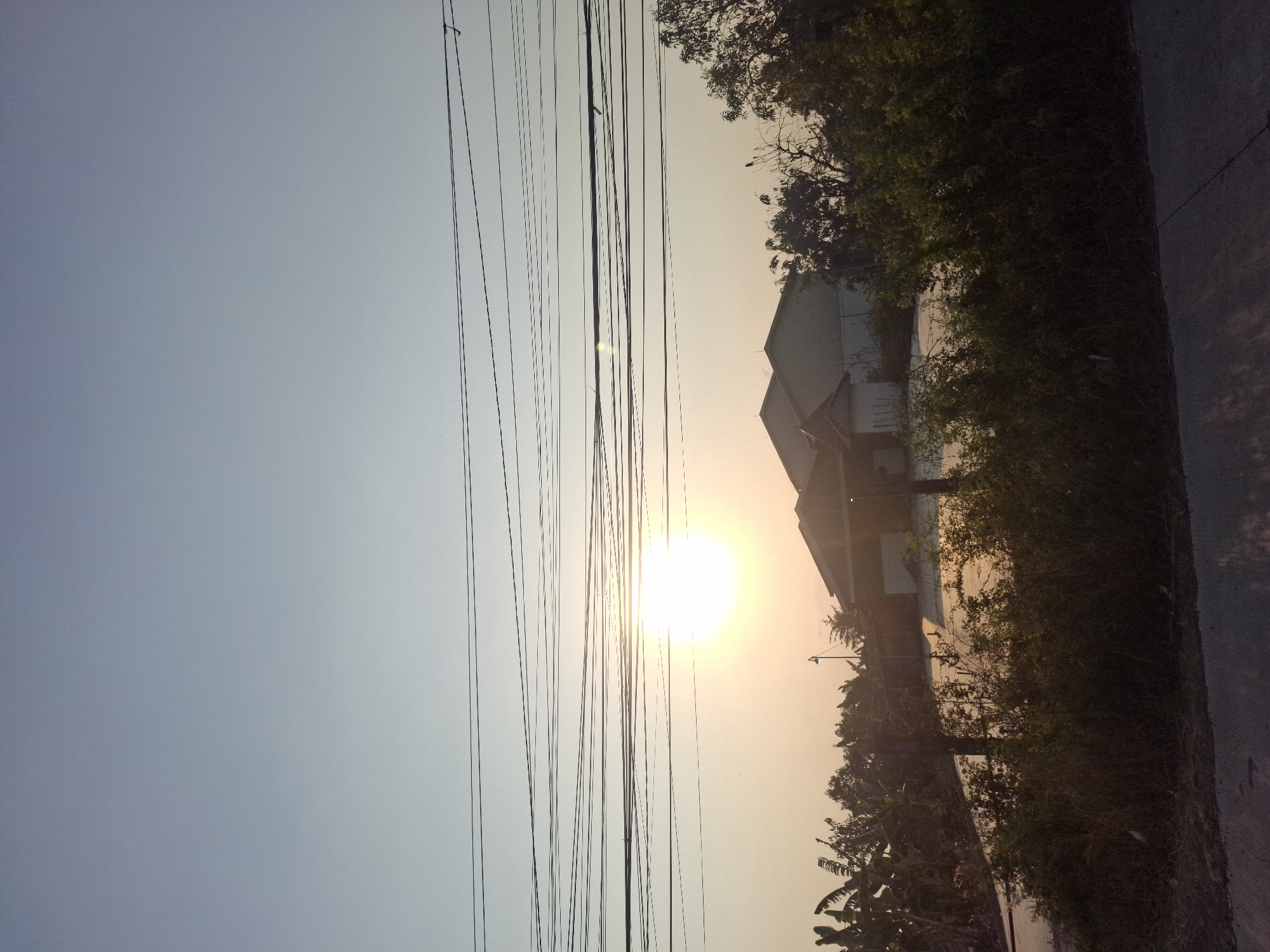 The image depicts an outdoor scene at sunset in Banjarmadu, Indonesia. The sky is a pale blue, fading to a soft orange near the horizon. A bright sun is visible, partially obscured by a dense network of overhead power lines. In the middle ground, two buildings with light-colored roofs are nestled amongst lush green trees and banana plants. A few streetlights are visible in the distance. The foreground features a dirt road and overgrown vegetation.
