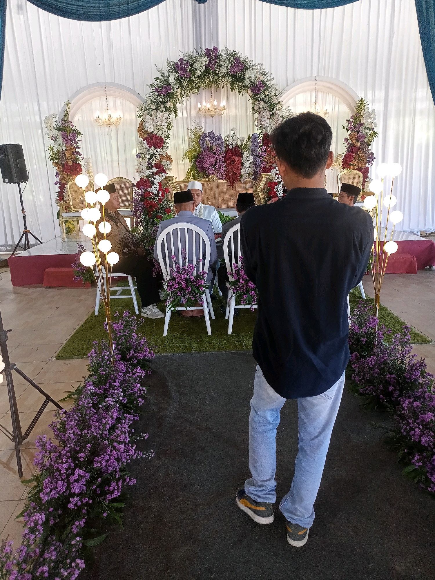 This media file captures a wedding ceremony in Wanadadi, Indonesia. The scene is set indoors, likely in a hall or tent, with white drapes forming the backdrop. A large floral arch, adorned with white, purple, and red flowers, frames the central area where the ceremony is taking place. Several individuals are seated on ornate chairs, dressed in traditional attire. One person in the foreground, facing away from the camera, appears to be a guest or photographer, dressed in a black shirt and light blue jeans.
The lighting suggests it might be daytime, possibly with artificial lighting supplementing. The overall atmosphere appears formal and celebratory, characteristic of a wedding. The presence of floral arrangements and decorative lighting indicates an effort to create a festive and aesthetically pleasing environment. The setting is richly decorated, with purple flowers lining the pathway to the seating area and flanking the floral arch, adding to the visual appeal. There is no visible text in the image.
