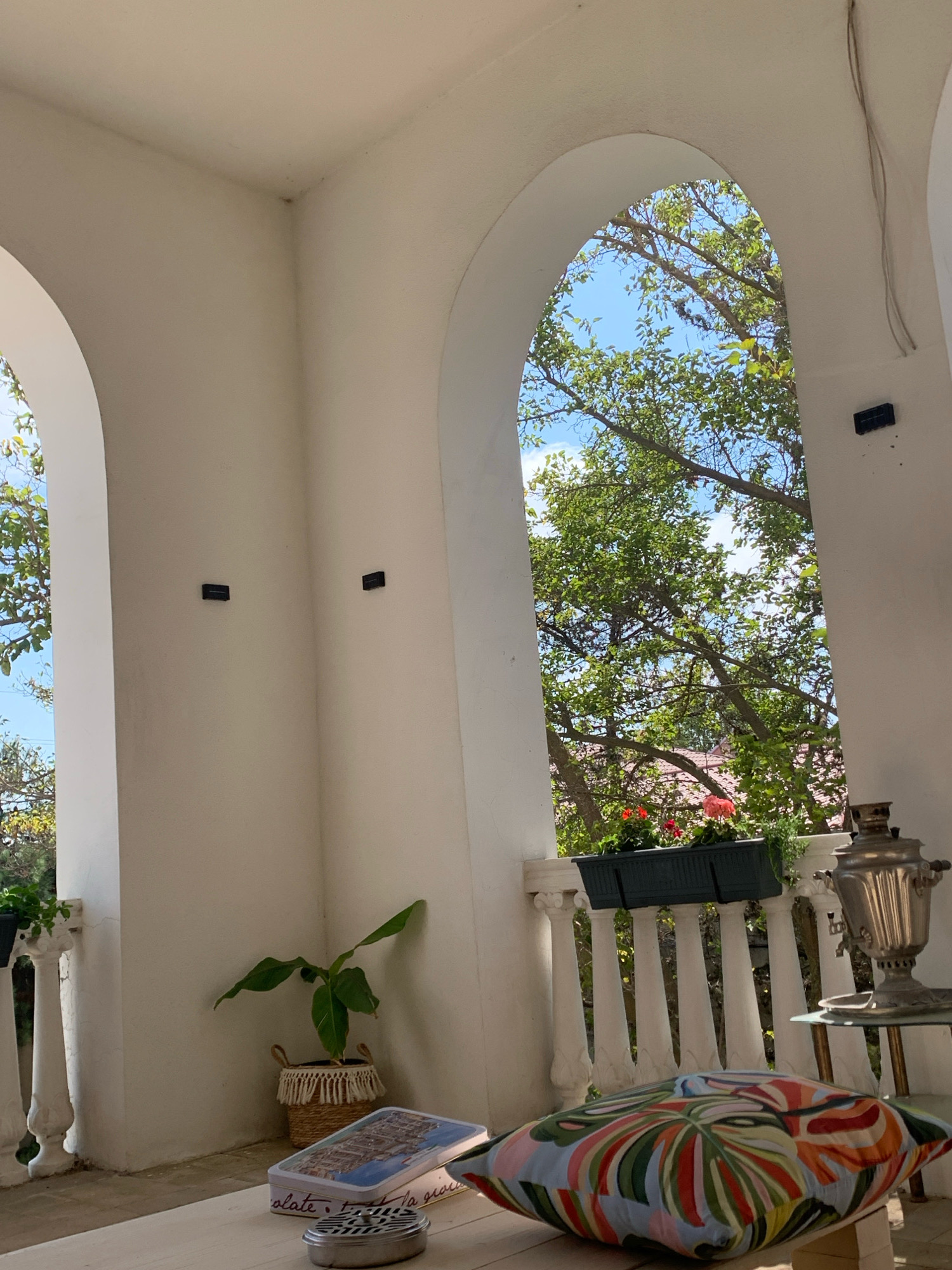 The image depicts a covered outdoor seating area, possibly a veranda or balcony, with architectural elements suggestive of a Mediterranean or Middle Eastern style, such as arched openings and white stucco walls. The scene appears to be set in a residential or guesthouse environment in Khazar Raion, Azerbaijan.
In the foreground, a large, vibrantly colored decorative pillow rests on a wooden surface, likely a floor or low table. To the left of the pillow is a rectangular metal tin with an image of buildings on it and a small, cylindrical metal object. Further left, a potted plant with large green leaves sits in a woven basket.
The midground reveals two large, rounded arches opening to an exterior view. Through the right arch, a balustrade with white, turned spindles is visible, supporting a row of dark green planters containing red and pink flowers. Beyond the balustrade, lush green trees with dappled sunlight filtering through their leaves and a bright blue sky are visible. A traditional Russian samovar sits on a small, round glass-topped table to the right of the arch.
The left arch provides a partial view of a similar balustrade and more greenery. The white stucco walls of the structure are visible throughout the image, with two small, dark rectangular objects mounted on the wall behind the right arch, possibly outdoor light fixtures or electrical outlets. The lighting suggests it is daytime, with sunlight casting shadows and highlighting the textures of the surfaces. No people are present in the scene.
