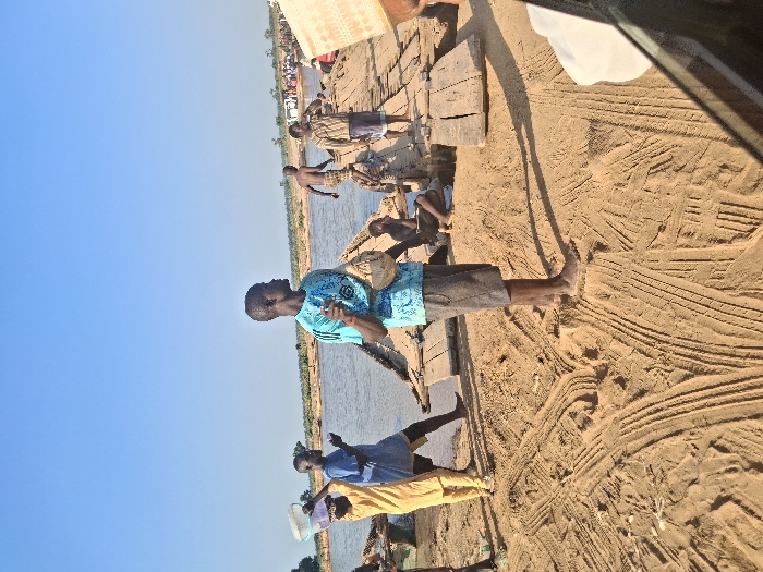The image depicts an outdoor scene at a river crossing in Nemnei, Nigeria, under a clear blue sky. The foreground consists of light brown sand exhibiting numerous tire tracks. In the center, a dark-skinned male stands barefoot, wearing a light blue patterned short-sleeved shirt and dark shorts, holding a small light-colored pouch and gesturing with his right hand. To his left, a person in a yellow garment carries a white and a purple plastic bucket on their head, facing towards the water. Behind them, another dark-skinned male in a blue shirt stands near the water's edge.
A makeshift wooden bridge or dock, constructed from rough planks, extends from the sandy bank into a wide body of water occupying the mid-ground. Several individuals are present on and around this structure. A shirtless dark-skinned male is stepping onto the wooden structure from the water, while another dark-skinned male in a plaid shirt and wrapper stands further along the planks, holding an object to his mouth. A child or small individual sits on the sand adjacent to the wooden structure.
The distant riverbank features sparse vegetation and more indistinct figures, suggesting further activity. A portion of what appears to be a vehicle's frame and a woven object are visible in the bottom right corner. The scene is brightly lit by natural sunlight, casting shadows to the left.