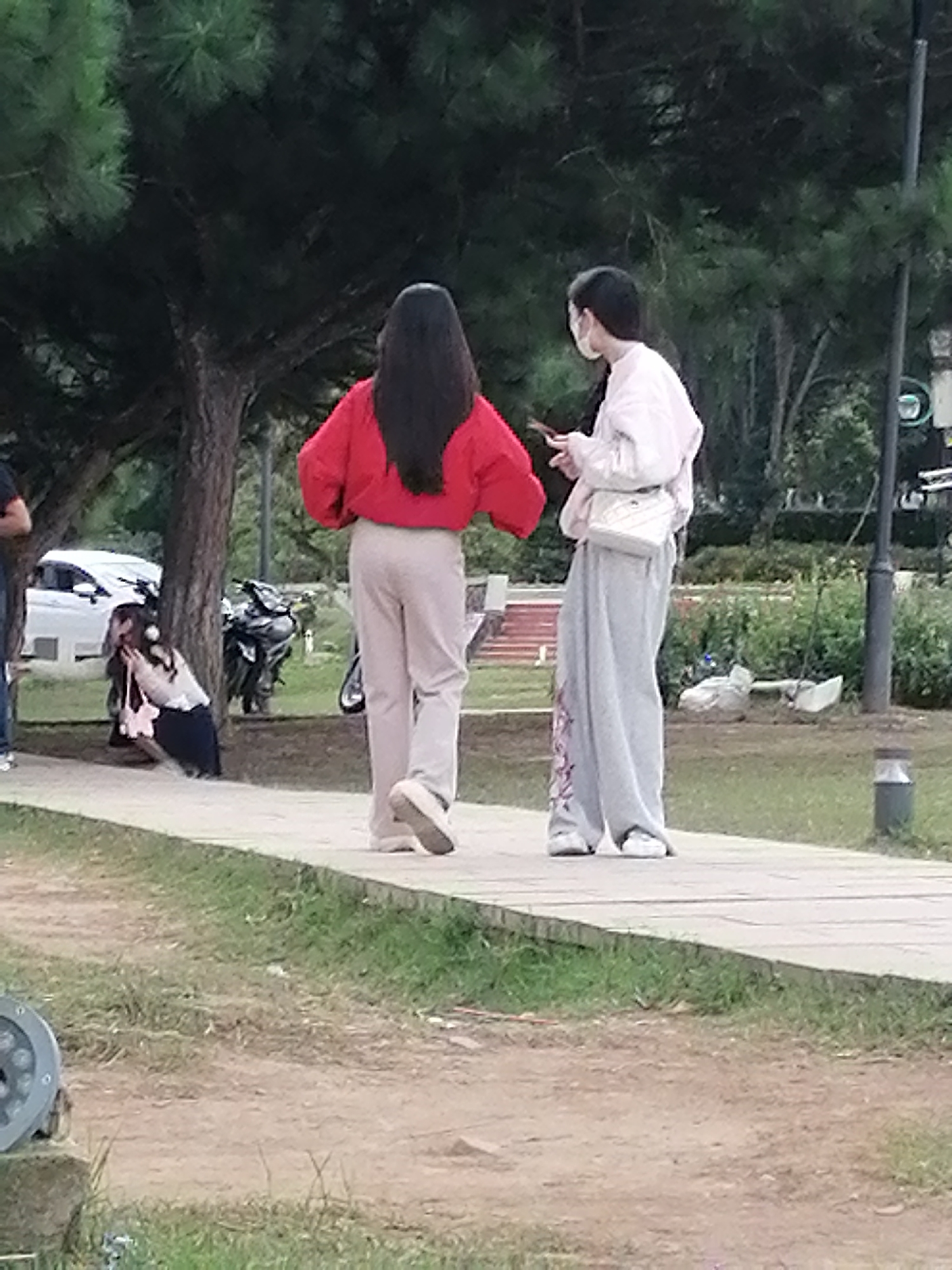 The media file shows two young women standing on a paved walkway in an outdoor park setting. The woman on the left has long, dark hair and is wearing a bright red sweatshirt and light-colored pants. The woman on the right is wearing a light-colored hoodie, a white crossbody bag, and loose-fitting gray pants with a pink pattern. She is looking down at her phone.
In the background, there are trees with lush green foliage, suggesting a warm or mild climate. A white car and a dark-colored motorcycle are parked behind the woman on the left, indicating this is an area where vehicles are permitted. Further in the background, there is more greenery and some visible structures, possibly part of the park or a nearby building.
To the left of the walkway, a third person, a young woman, is crouched down, appearing to be taking a photo or interacting with something on the ground. She is wearing a light-colored top and a dark skirt or pants. A pink bag is next to her.
The lighting suggests it is daytime, likely late afternoon or early evening, as the light is diffused and there are shadows. The overall atmosphere appears relaxed and casual. The location cues, such as the foliage and potentially the style of the park infrastructure, are consistent with a place like Da Lat, Vietnam. There is no visible text in the image.