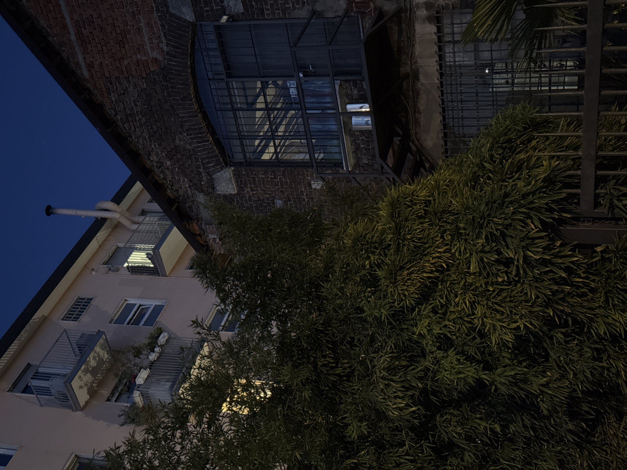 Captured at night, the image presents a view of a building complex, likely in Milan, Italy. A multi-story residential structure, painted in a light hue, stands to the left, with a series of balconies and windows, some of which are illuminated. Adjacent to it, a taller brick building is visible, topped with a metal frame structure. Dense foliage dominates the foreground and the right side of the frame, partially obscuring the buildings. Above, a dark blue sky is apparent. A fence runs vertically along the right side of the frame.