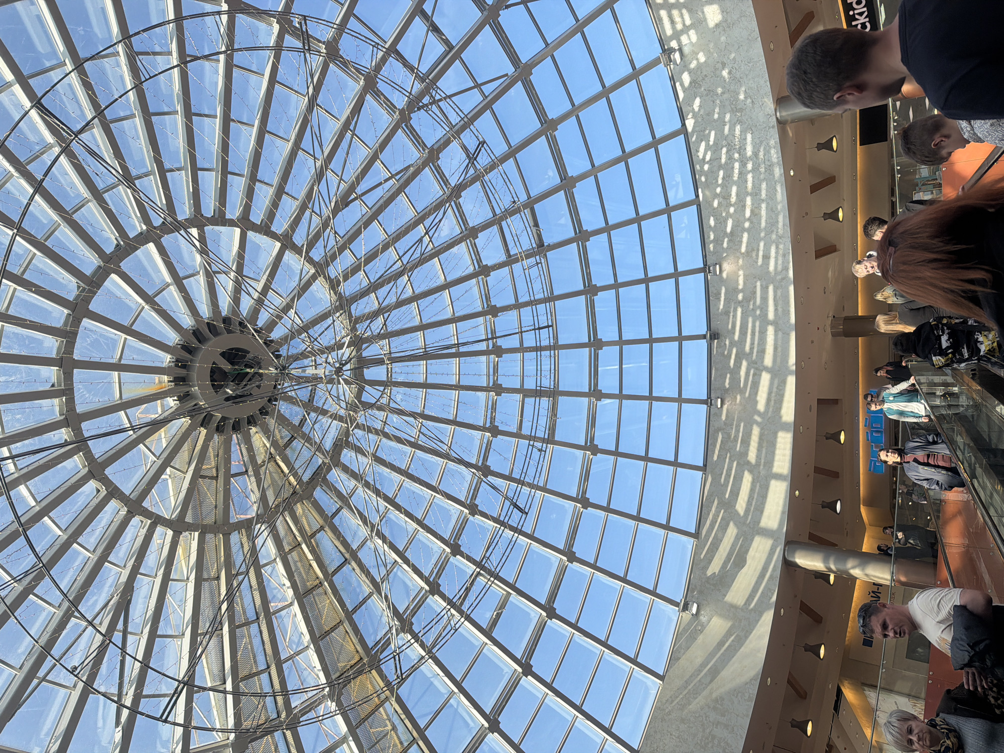 This image depicts the interior of the TRC Semya shopping mall in Perm, Russia, specifically focusing on its central atrium and domed skylight. The perspective is looking upwards from a lower level.
The primary architectural feature is a large, translucent glass dome forming the ceiling, supported by a radial metal framework. This framework consists of numerous light-colored rectangular and trapezoidal panels. A circular central hub is visible at the dome's apex. Thin wires strung with small, numerous lights, possibly decorative or functional, are suspended across the interior surface of the dome.
Below the dome, the curved walls and ceiling of the mall's upper levels are visible. These surfaces are light-colored, possibly concrete, with a textured finish. Recessed lighting fixtures and suspended pendant lights are integrated into the ceiling. Bright natural sunlight streams through the dome, casting distinct grid-like shadow patterns onto the upper wall surfaces. A cylindrical support column is located on the left side of the scene.
Two parallel escalators ascend diagonally from the foreground to the midground, featuring glass and metal railings. Approximately 20-30 individuals are visible on these escalators and on the surrounding levels. They are dressed in casual attire, consistent with shoppers or mall visitors.
In the left foreground, a woman with short, light-colored hair and a dark patterned scarf stands beside a man in a white t-shirt with sunglasses positioned on his head. He holds a dark object. In the right foreground, the backs of two individuals and a child's head are visible. Various other people are distributed along the escalators and on the visible mall levels, primarily engaged in movement.
Partial Cyrillic signage "АЙ-П" is discernible on a wall to the left, and a partial English sign "ckid" is visible on the right, indicating commercial establishments within the mall. The scene is brightly lit by natural light from above.