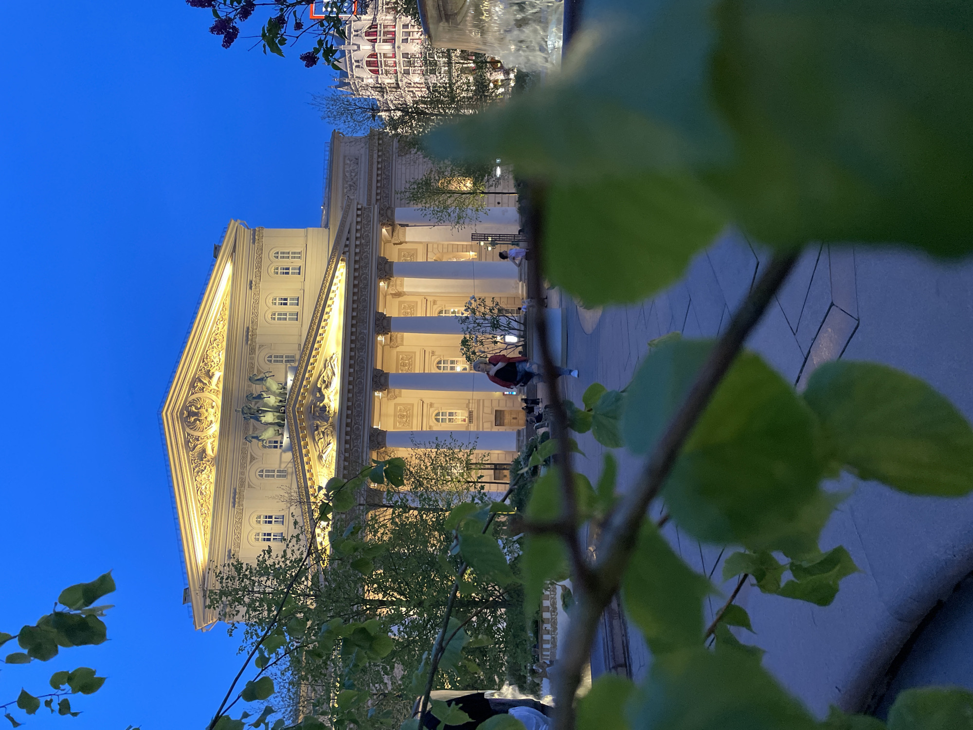 The Bolshoi Theatre, a historical landmark in Moscow, Russia, is captured in an upward angle shot. The theatre's facade is illuminated against the dark blue evening sky. Its columns are visible, as well as a sculpture or relief above the entrance. People are seen on the steps of the theater. In the foreground, a branch with green leaves is partially obscuring the view. A building or structure can be seen across from the theater in the background. The overall environment appears to be an urban setting at dusk or night.