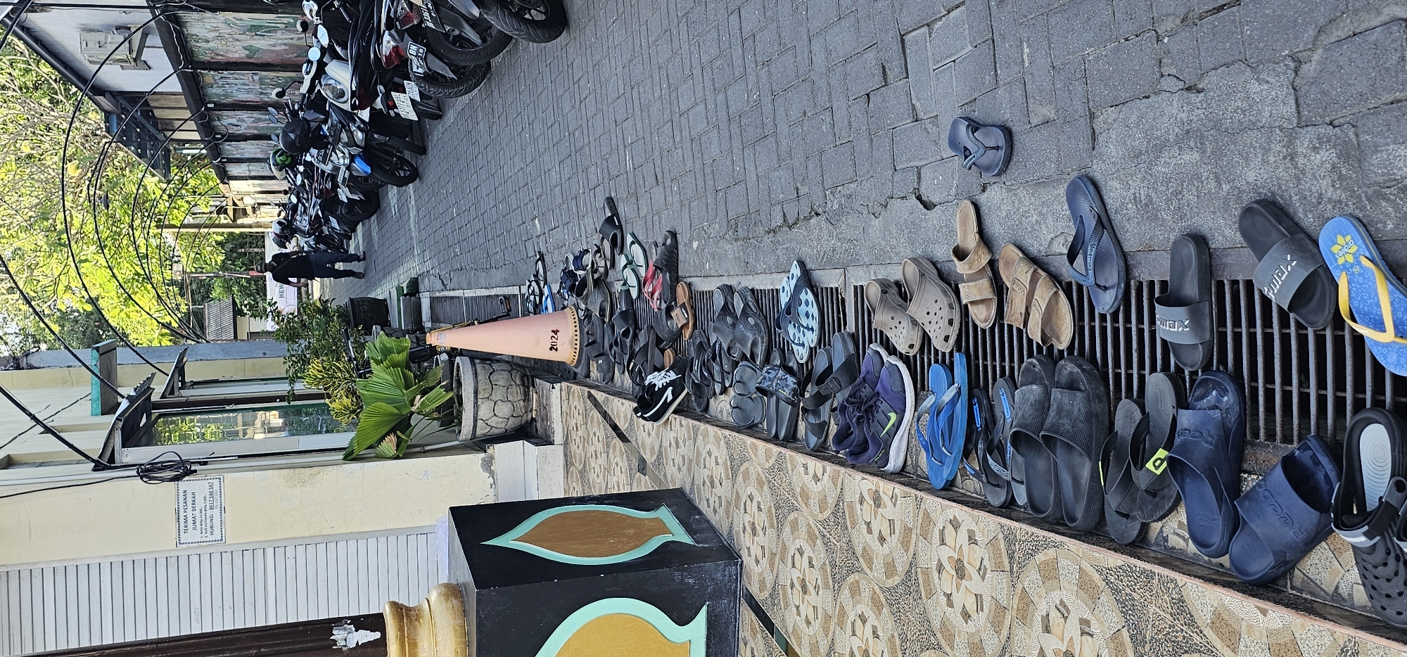 This outdoor scene in Denpasar, Indonesia, shows numerous parked motorcycles in the background, lining a gray-bricked walkway. In the foreground, an array of shoes and sandals of various styles and colors are arranged along a decorative patterned ledge adjacent to a metal grate. A traffic cone with "2024" written on it lies on the ground amongst the shoes. A black box with ornamental details sits near the ledge, along with a potted plant. Further left, a building's facade is visible with a sign on the wall and louvered shutters below. Two people are walking near the parked motorcycles in the background.
